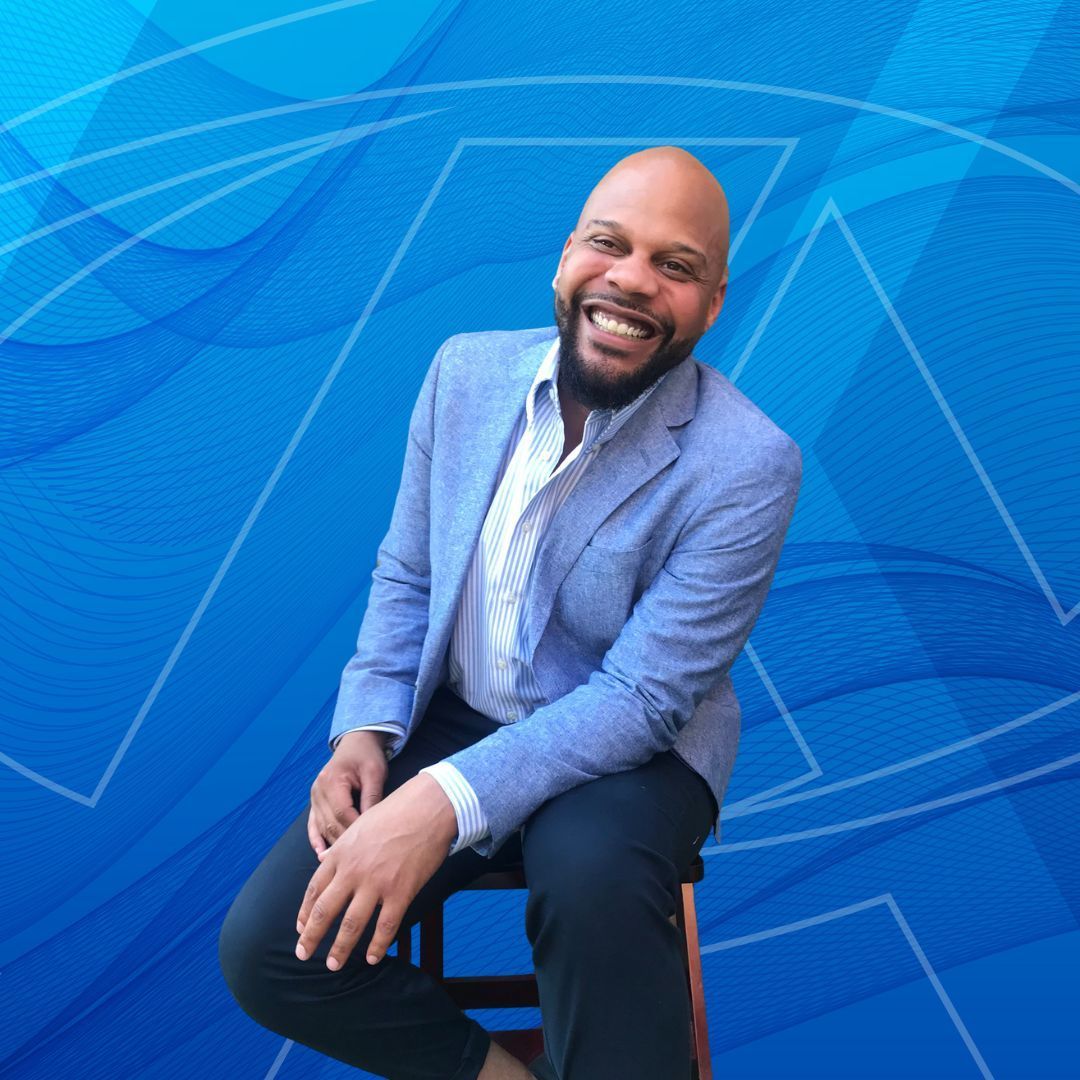 A bald man with a beard is sitting on a stool in front of a blue background.