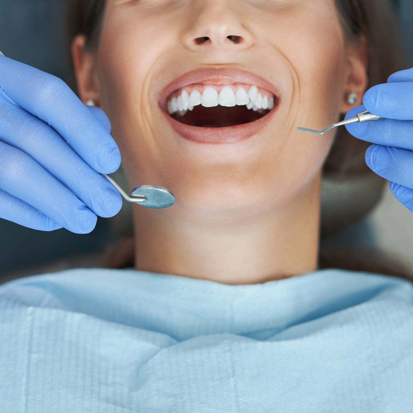 Woman at dentist's office with open mouth; dentist holding tools, wearing blue gloves.