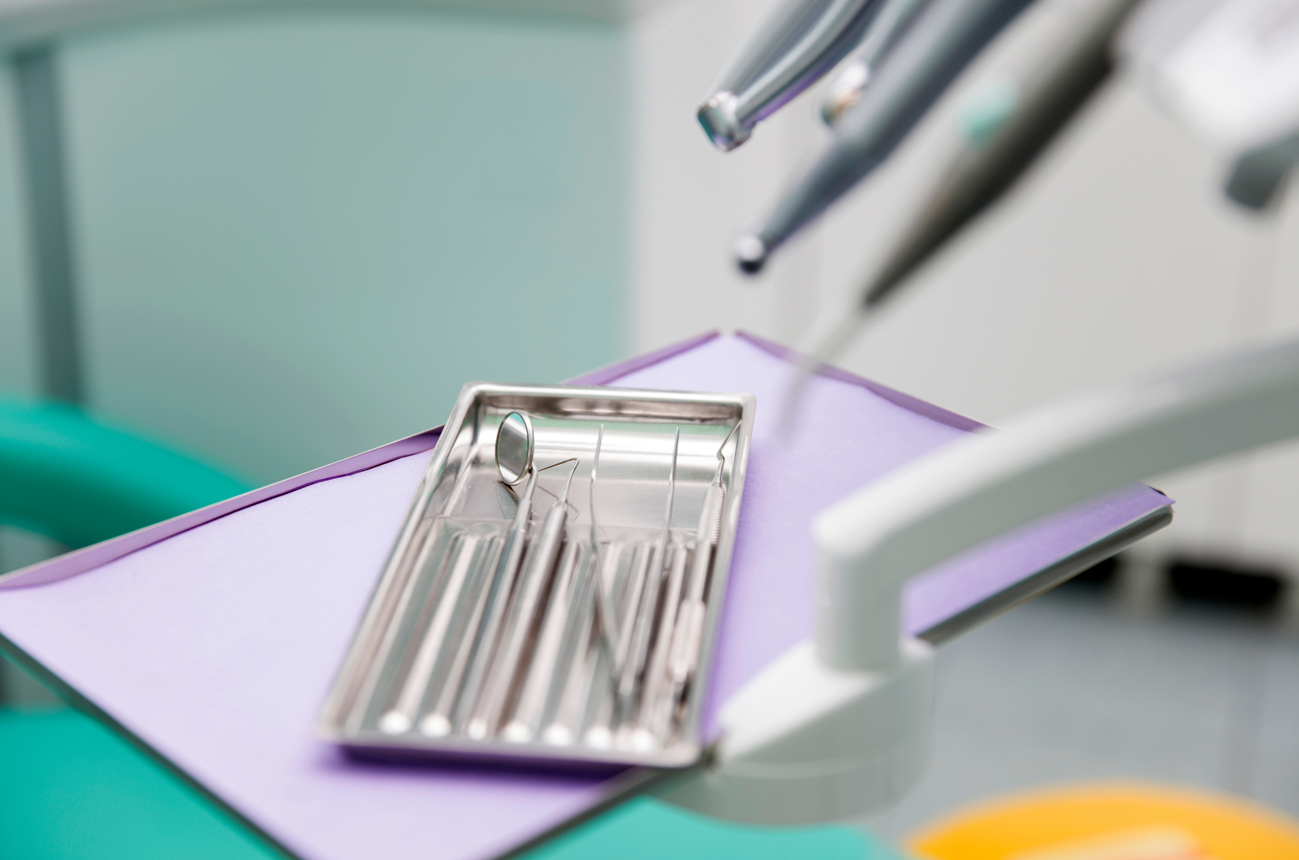 Dental tools in a metal tray on a purple drape, with dental chair and equipment in a clinic.