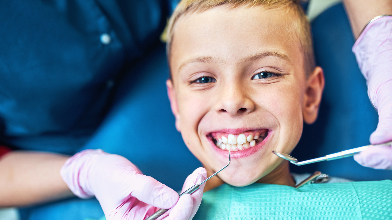 Boy in a dentist's chair smiling with mouth open; dental tools and gloved hands are visible.