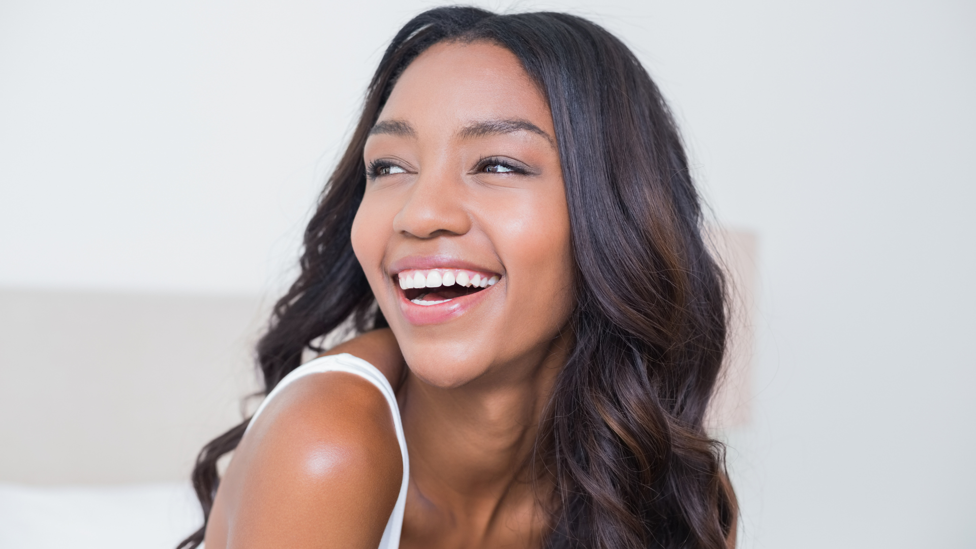 Woman with long dark hair smiles, looking off to the side, in a light-colored room.