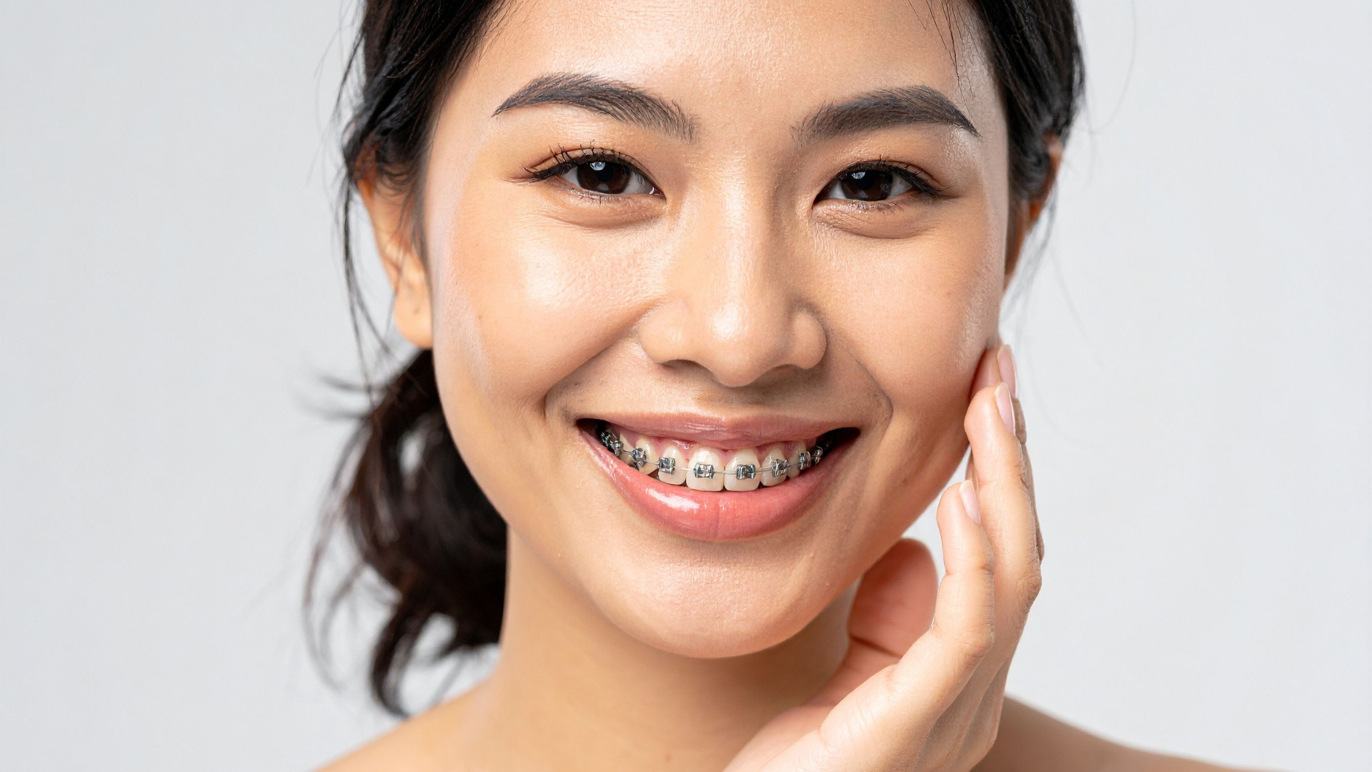 Smiling person with braces, touching cheek, against a light gray background.