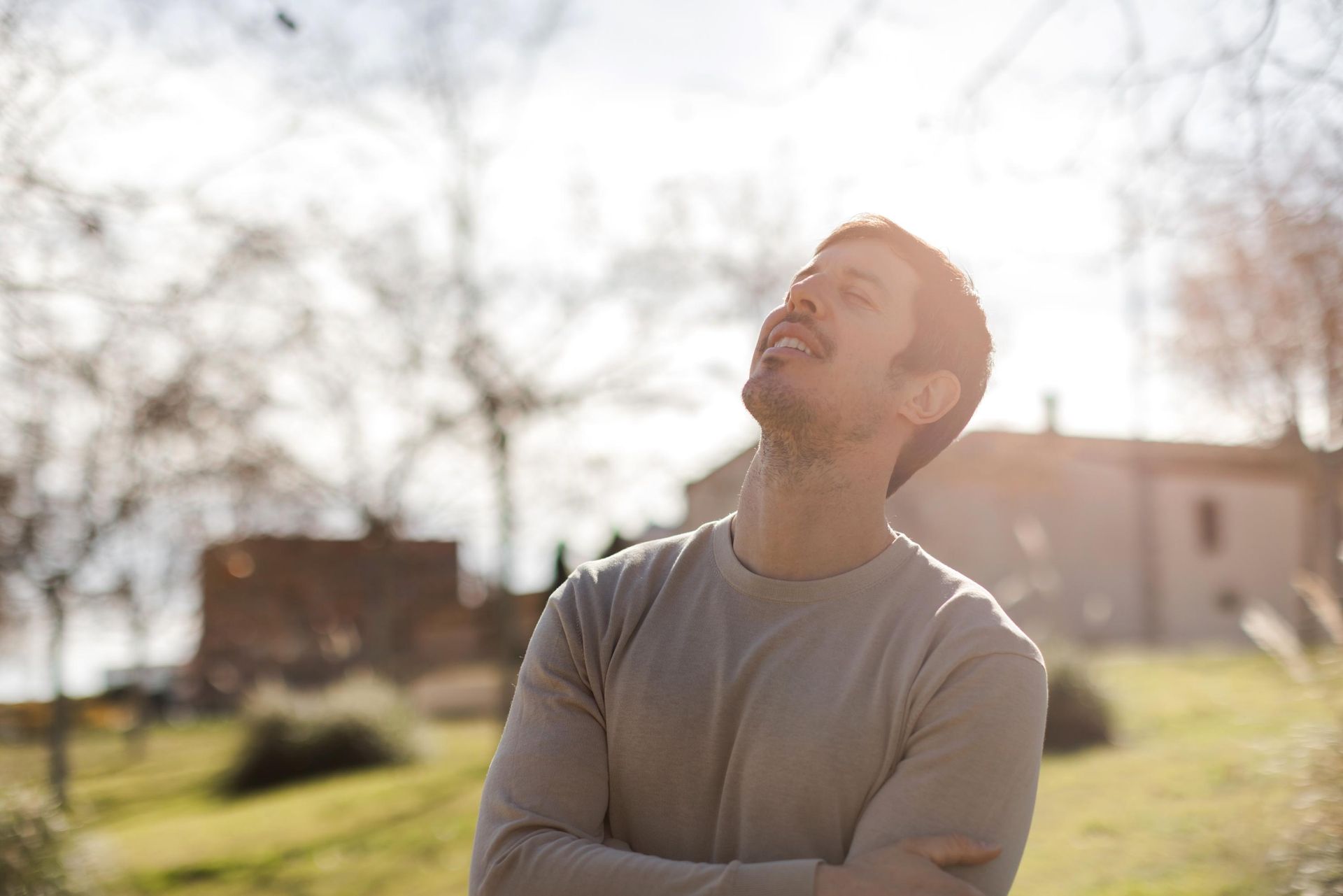 Man looking up at the sun with closed eyes, smiling. Outdoors, sunlight shining, trees and building blurred background.