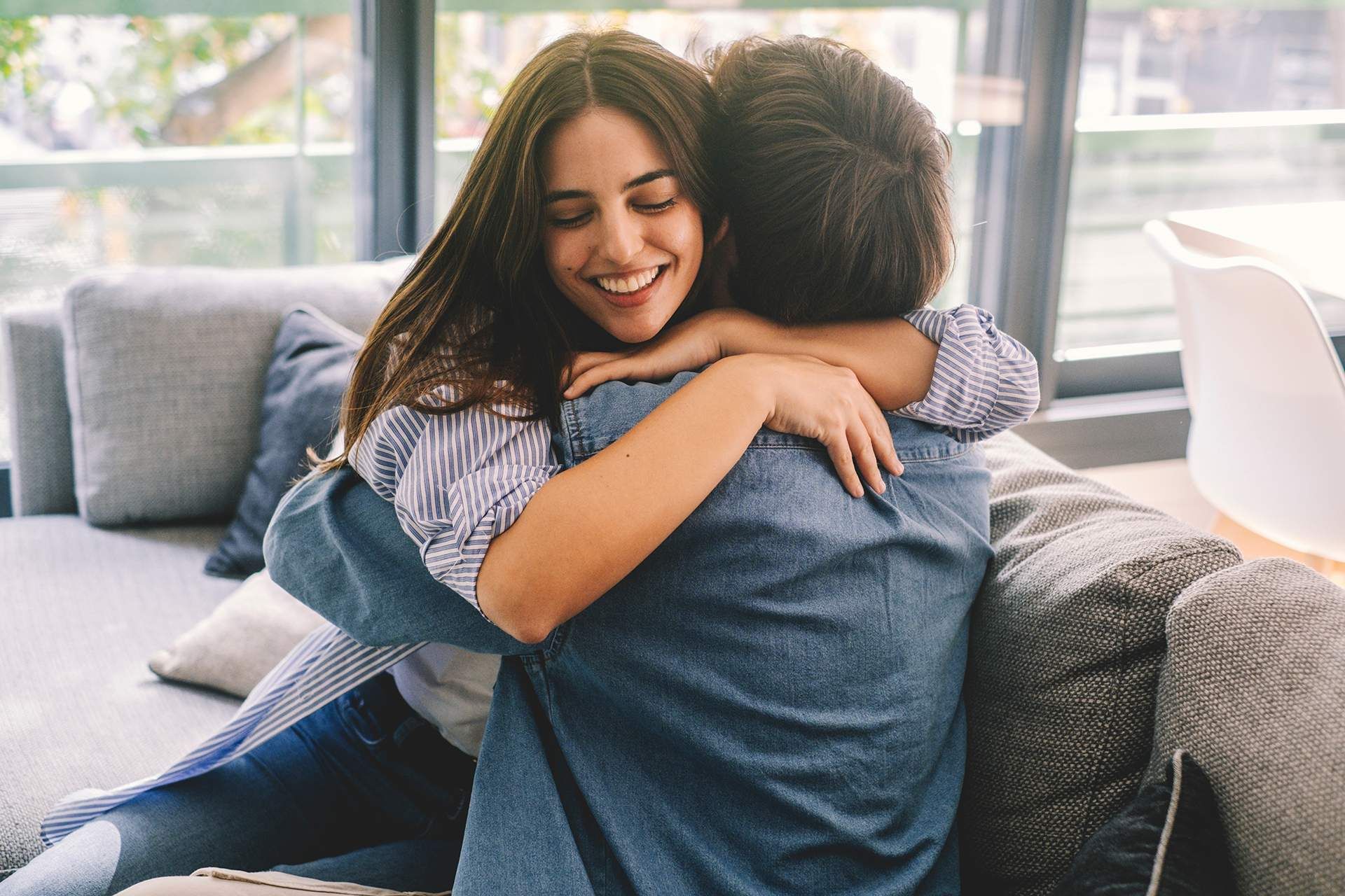 Woman smiling, embracing a person on a sofa indoors. Sunlight streams through a window.