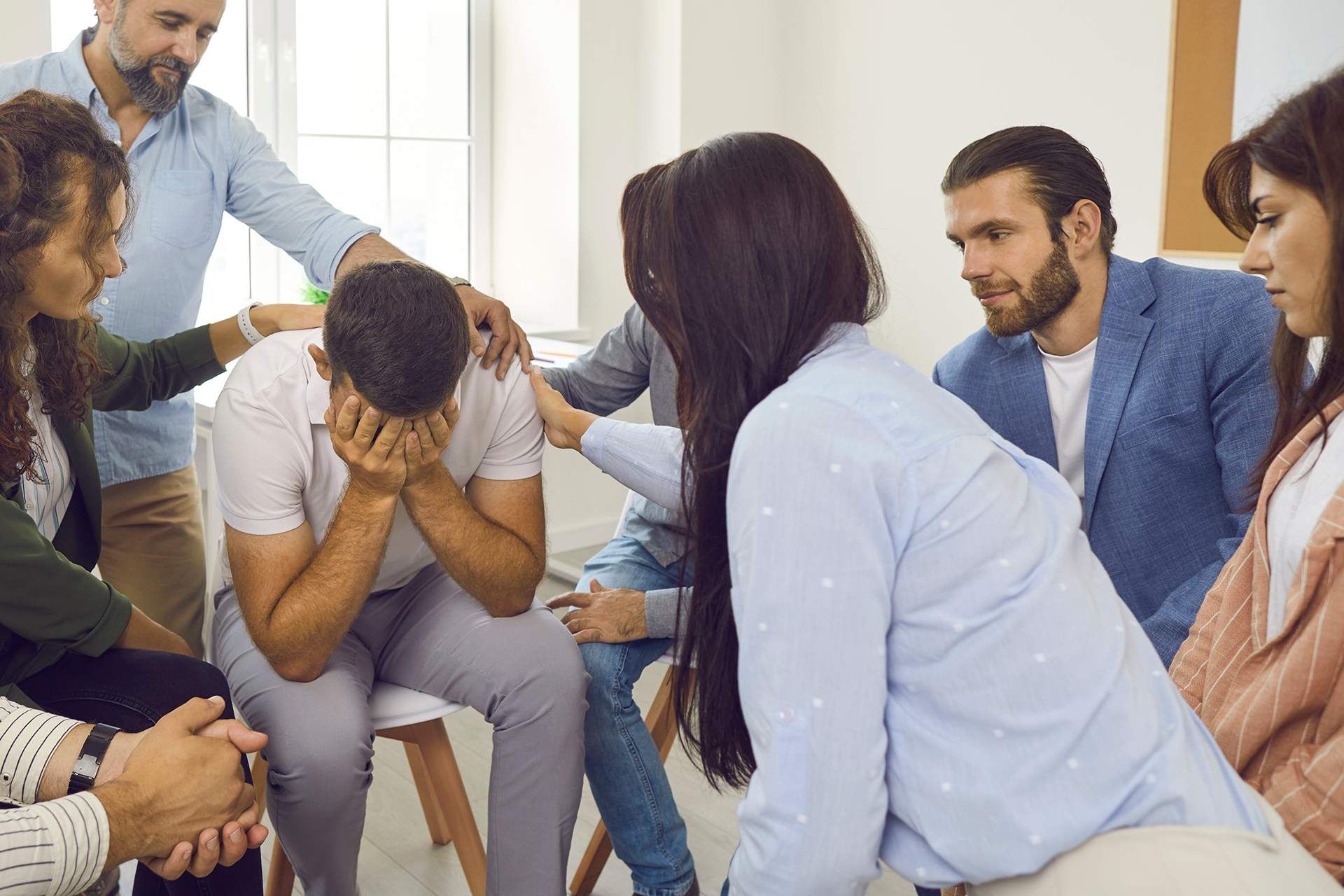 Group therapy session: Man cries with support from others in a circle, arms around him.