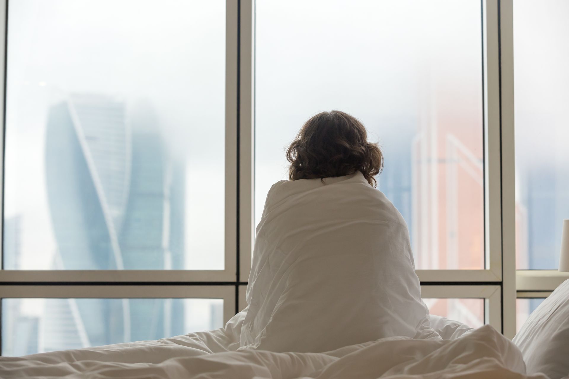 Person sitting on a bed by large windows, looking out at a hazy city skyline.