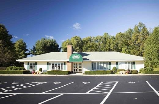A white house with a green awning is surrounded by trees and a parking lot.