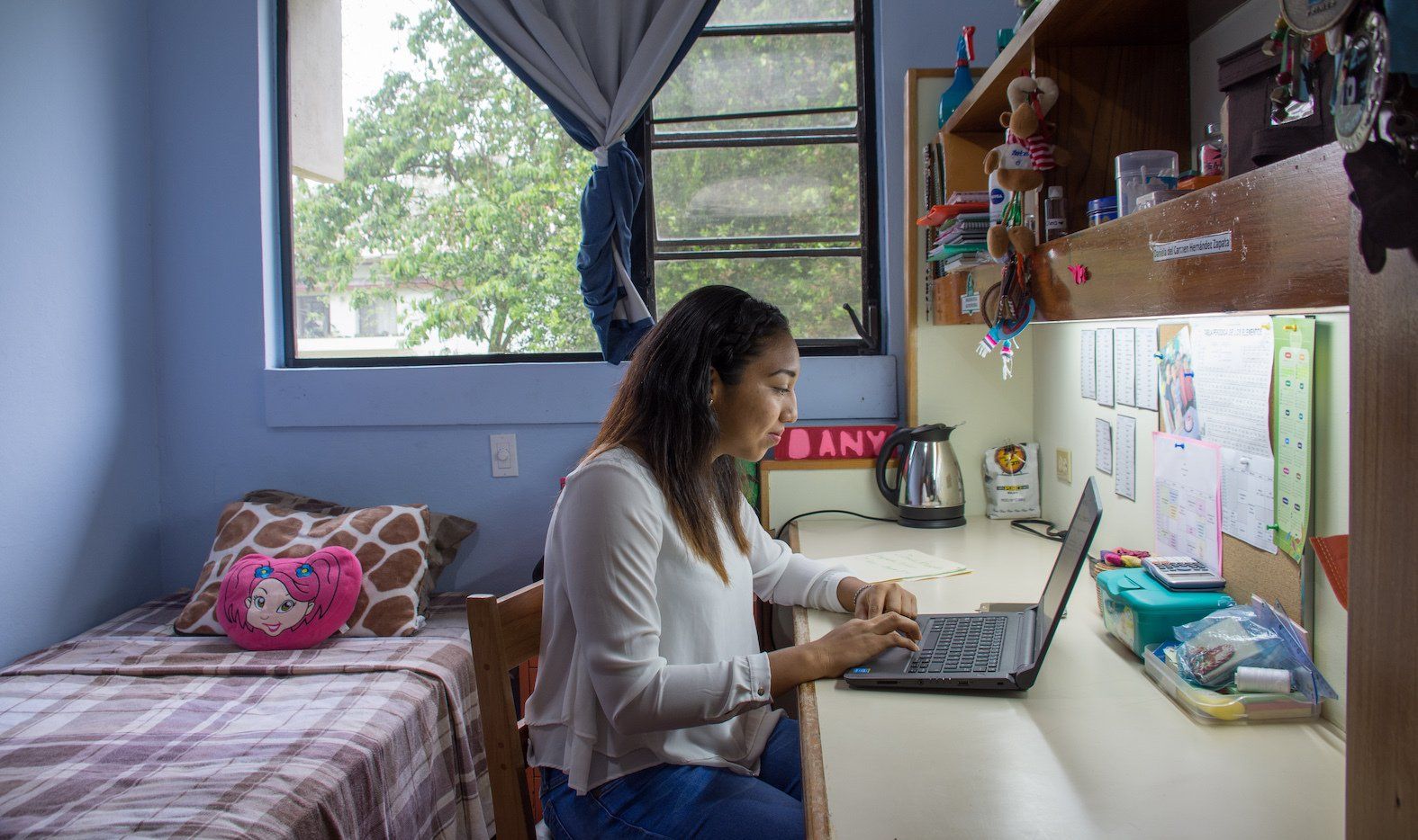 A woman is sitting at a desk in a dorm room using a laptop computer.