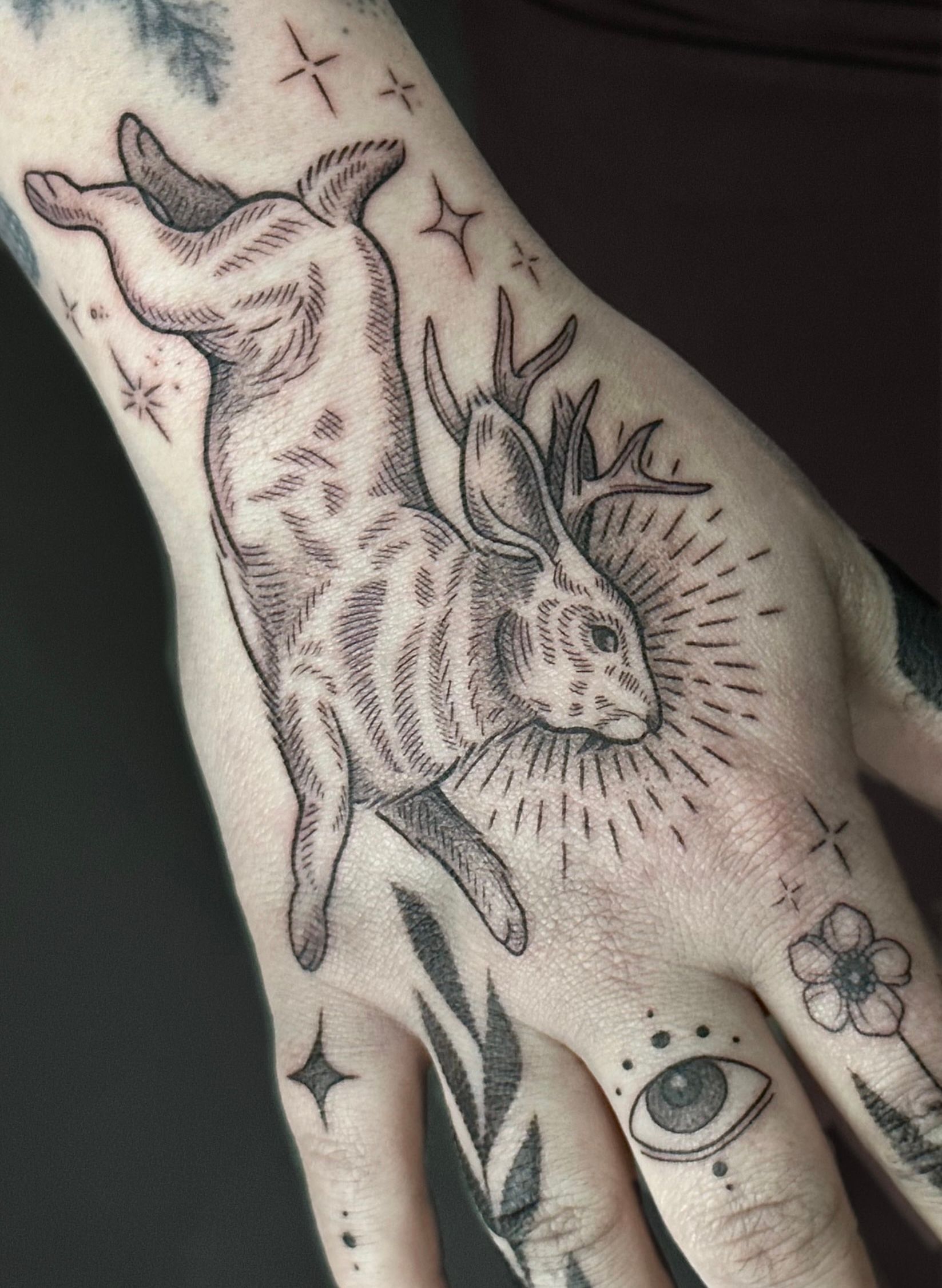 Hand tattoo: spotted rabbit with antlers and sparkling halo. Other tattoos: eye, floral, leaves, and stars.