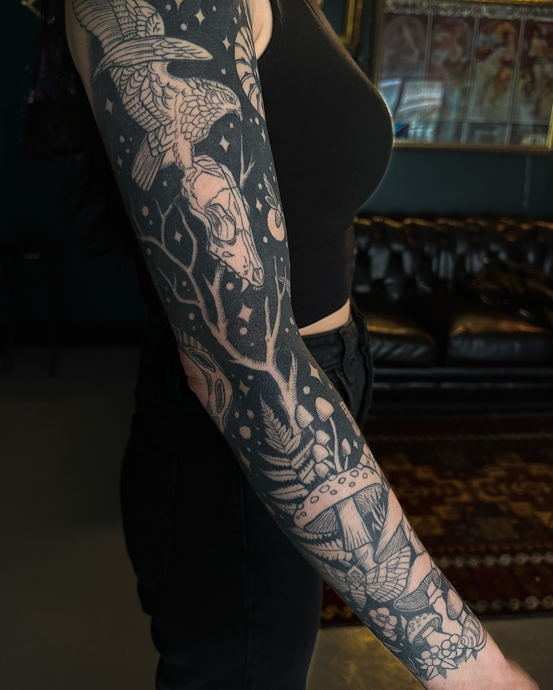 Person with a full-sleeve black tattoo on arm, wearing black top and pants, standing indoors.