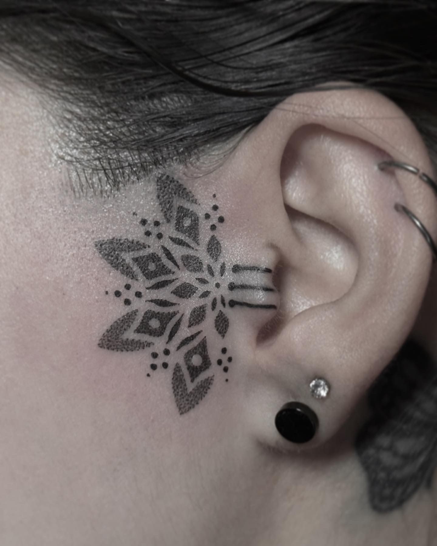 Ear with geometric black tattoo near ear and ear piercings.