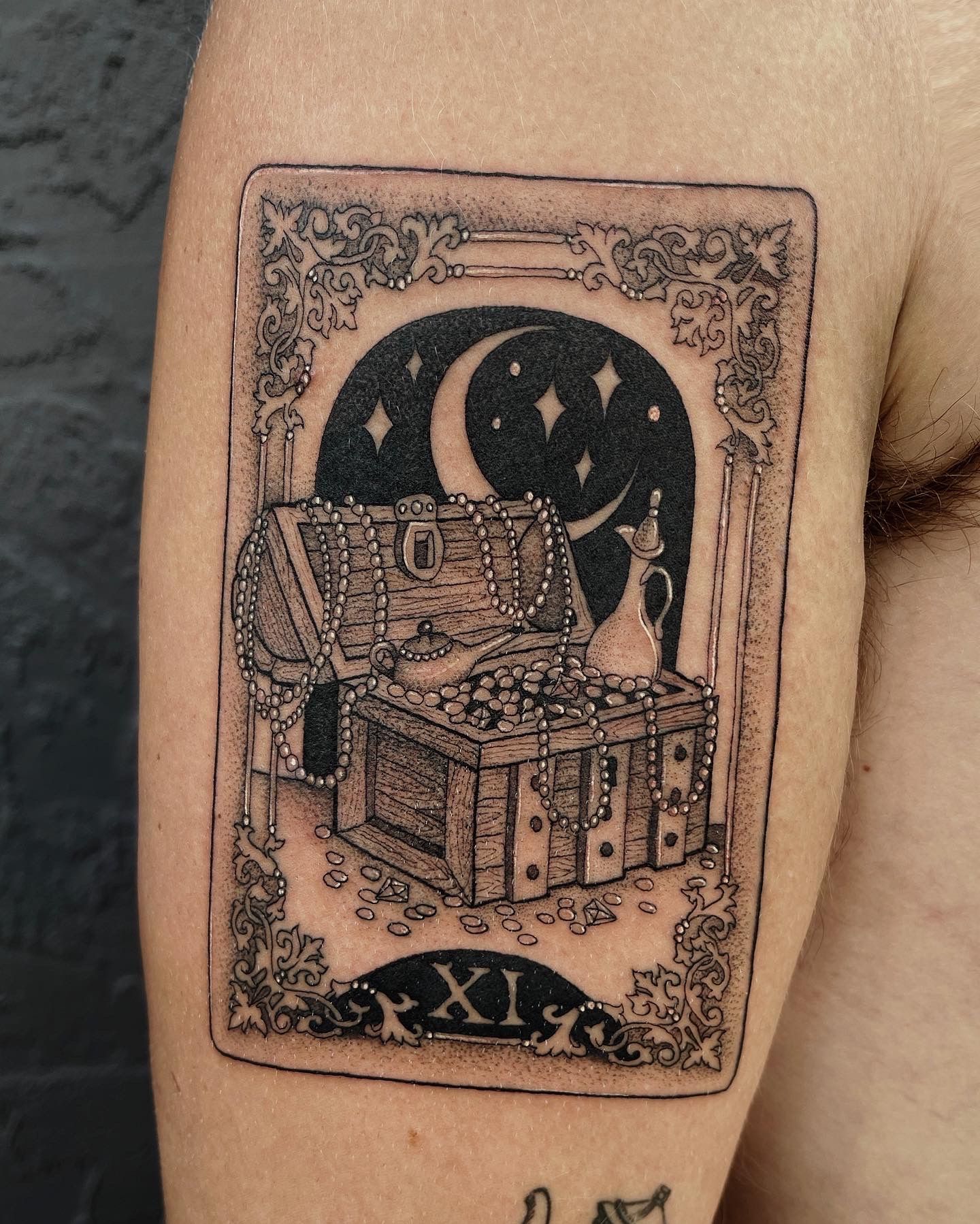 Tattoo of a black and white tarot card. Features a treasure chest under a crescent moon and stars, labeled with the roman numeral 