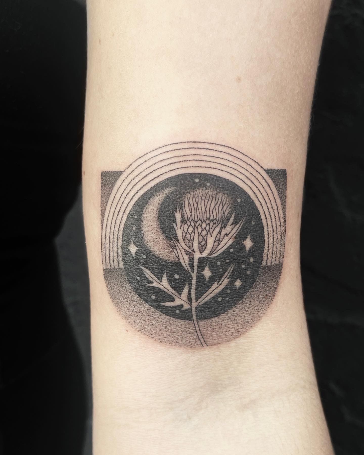 Tattoo of a thistle within a starry circle, with a crescent moon and arching lines, on skin.