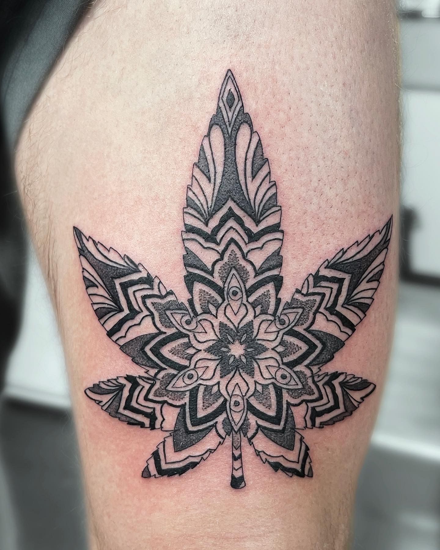 Black and grey cannabis leaf tattoo with mandala-style intricate patterns on the upper thigh.