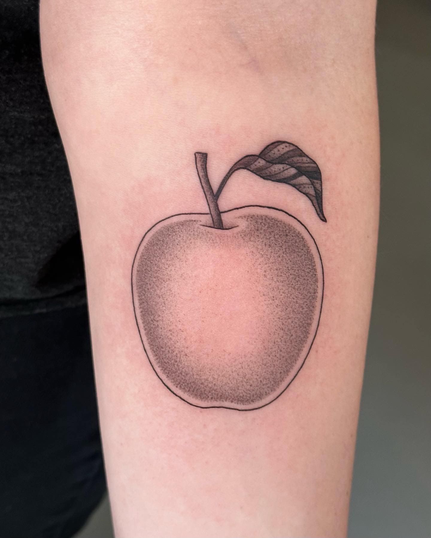 Tattoo of a shaded apple with a stem and leaf on a person's arm.