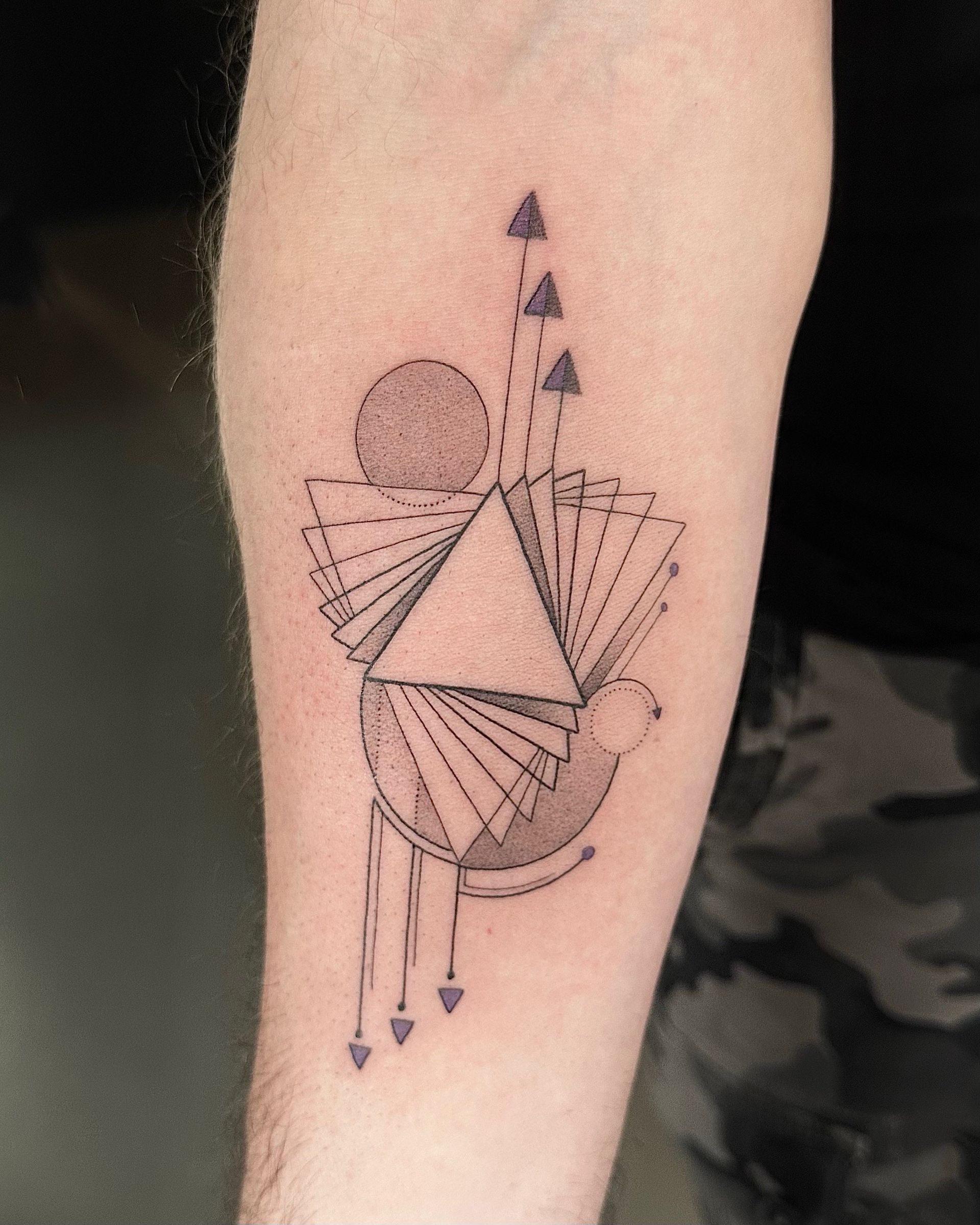 Tattoo of geometric shapes on an arm: triangle, circle, lines, and small triangles.