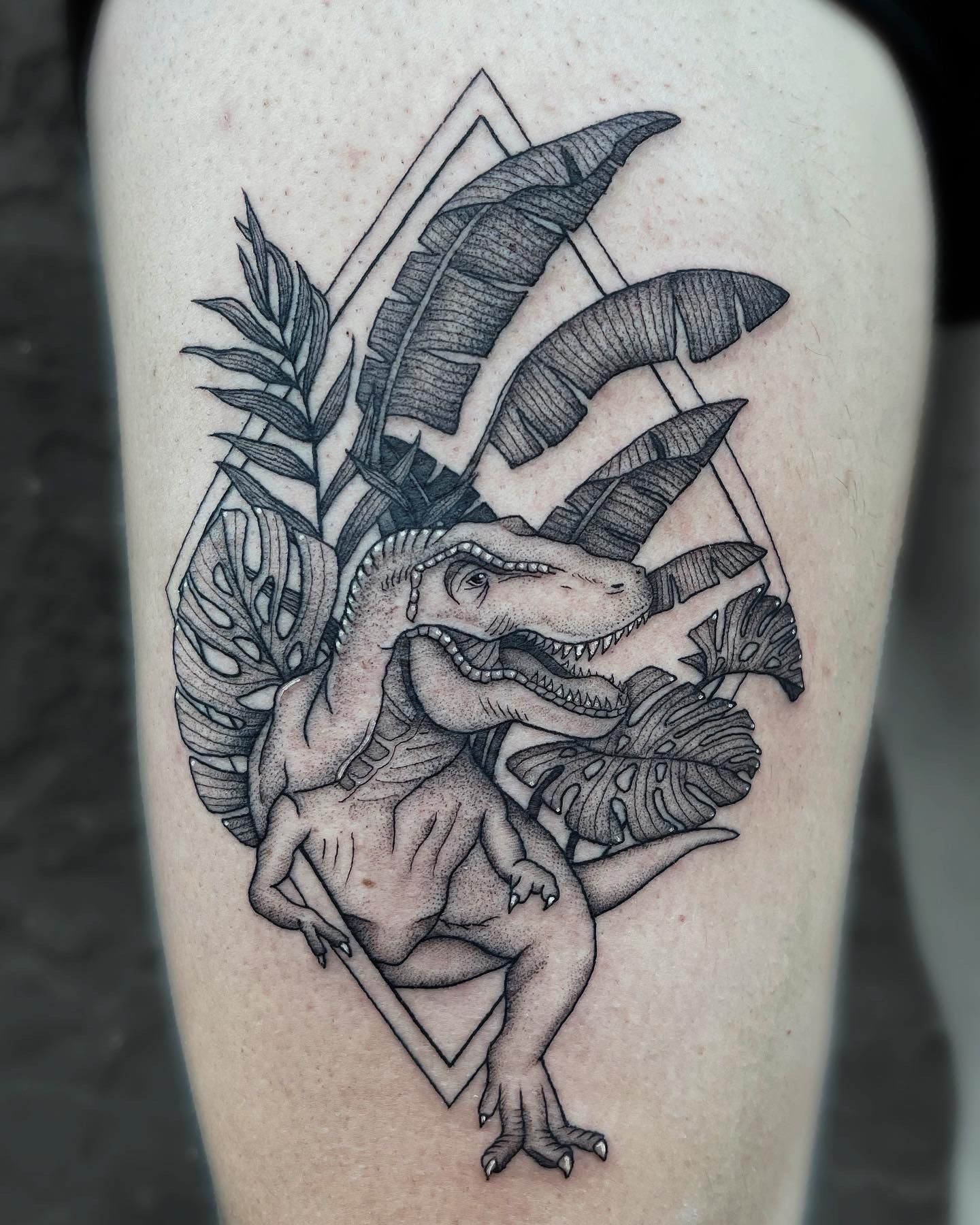 Tattoo of a Tyrannosaurus Rex surrounded by tropical leaves within a diamond outline.