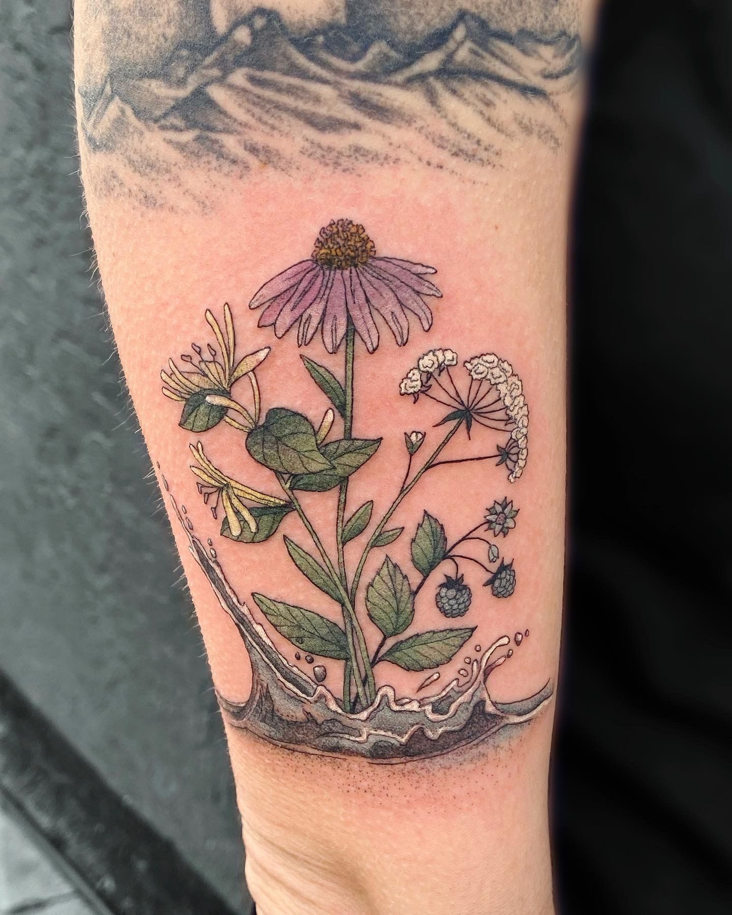 Tattoo of wildflowers with purple petals and green leaves, emerging from a splash of water, on an arm.