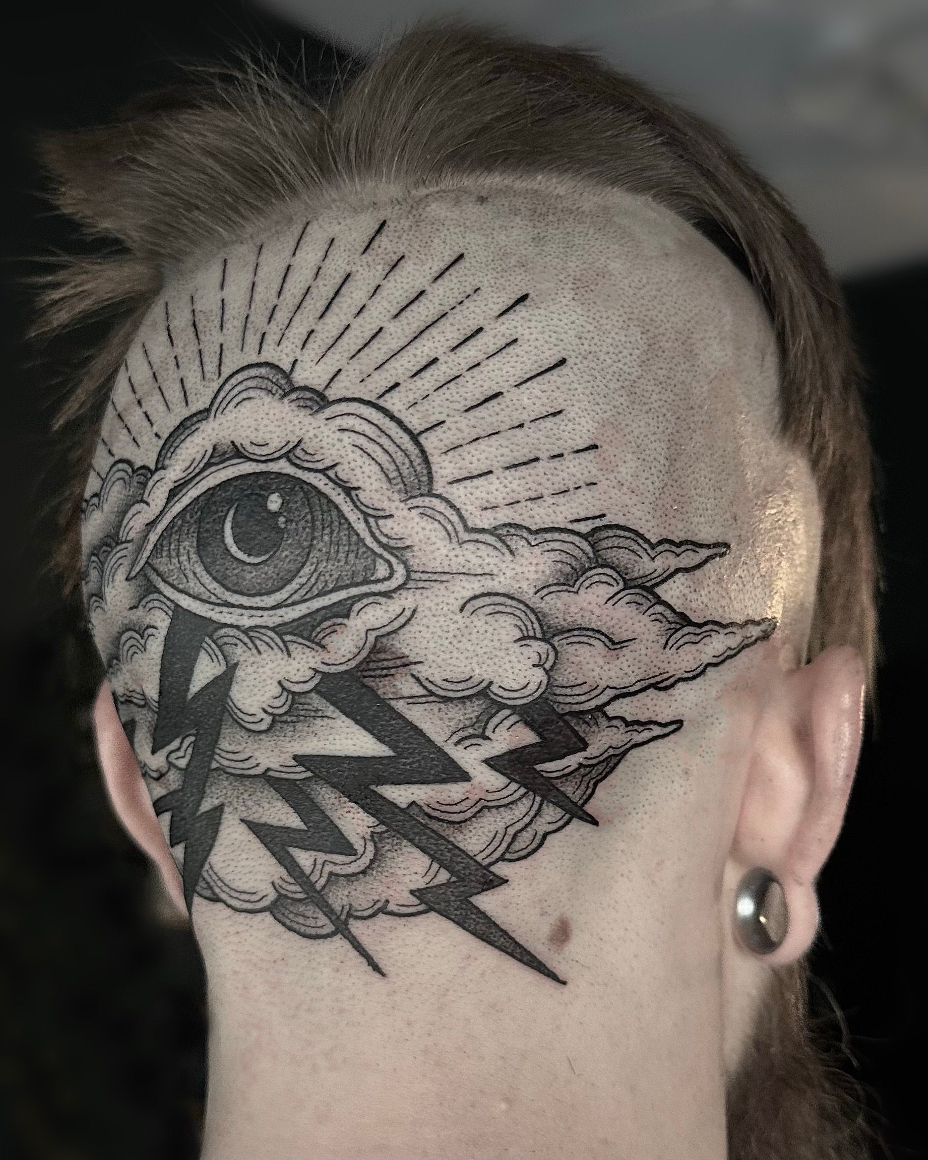 Person's head with a tattoo of an eye inside a cloud, sun rays, and lightning bolts.