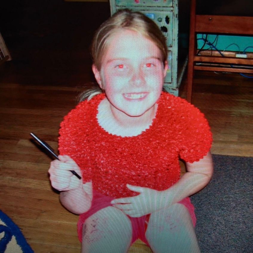 Girl in red shirt and shorts smiling, holding pen, seated on wooden floor.