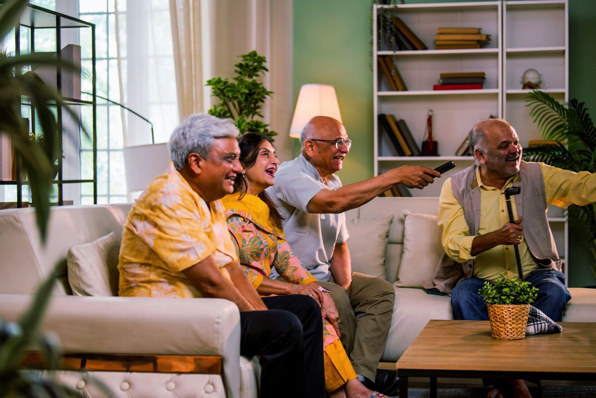 Four older adults on a couch watching TV, man holding remote, smiling, living room setting.