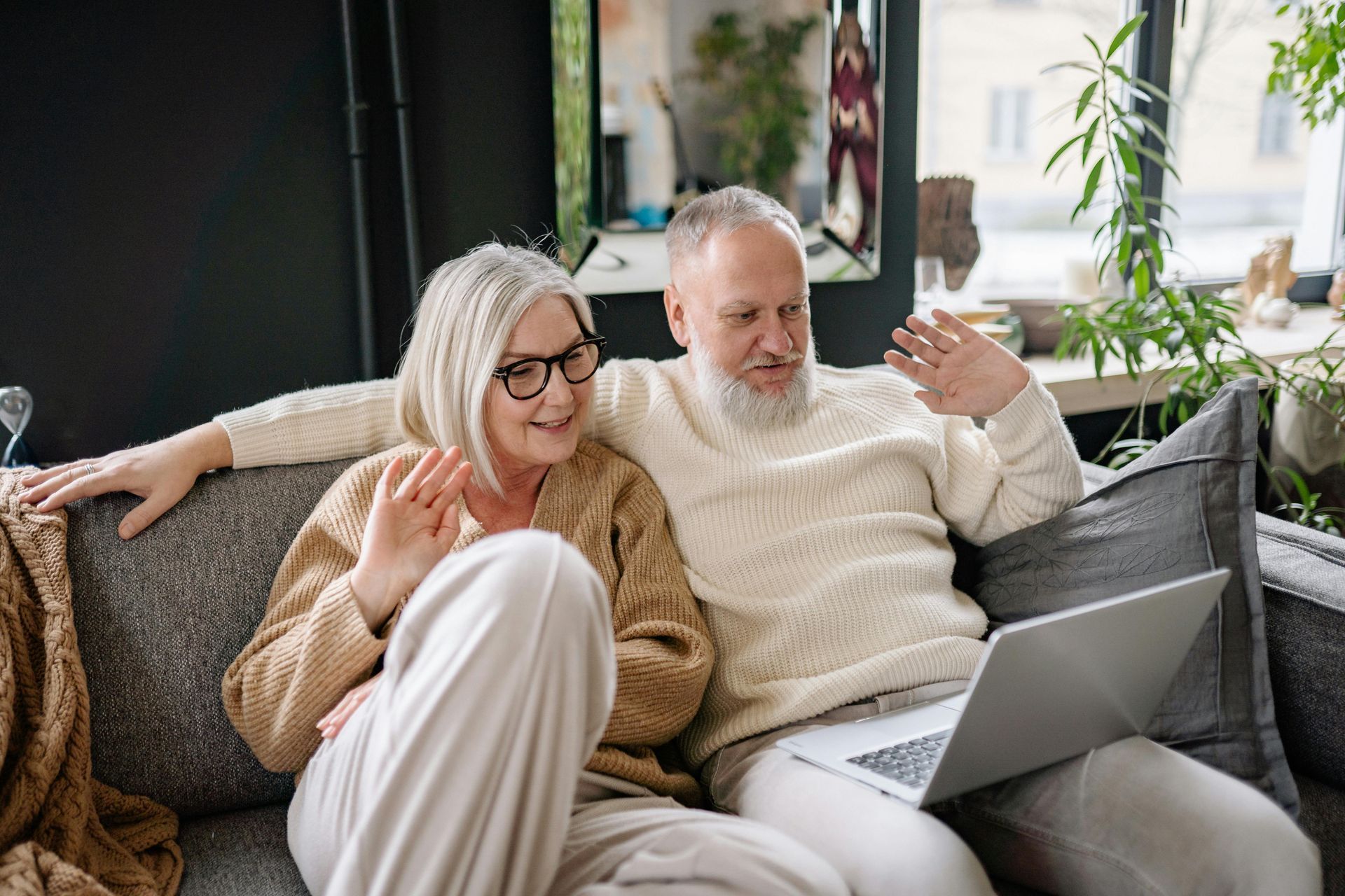 Elderly couple waving at laptop, sitting on a couch. Beige sweaters, glasses, neutral-colored room.