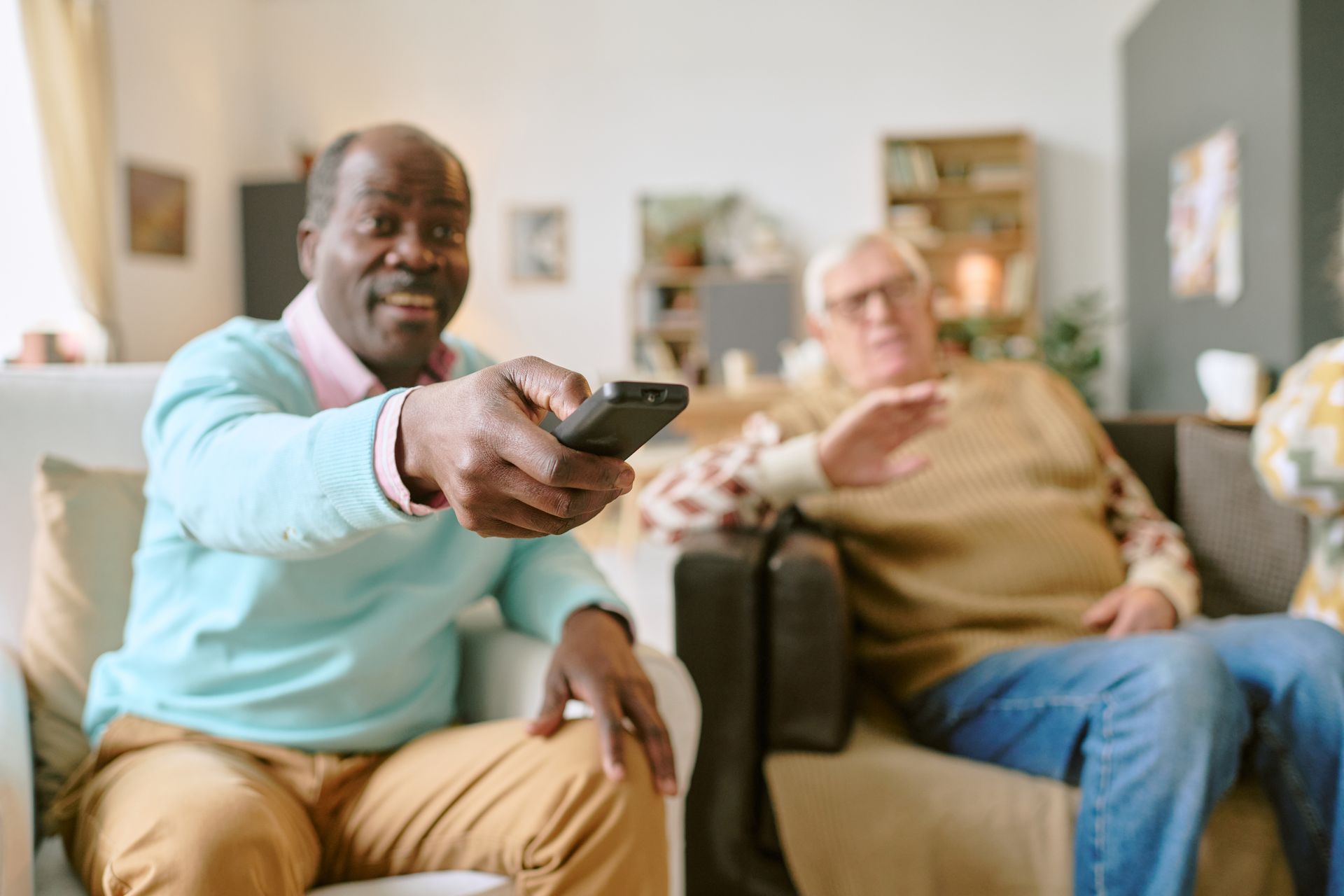 Two older men watching TV, one holding a remote with a smile, the other gesturing, in a living room.