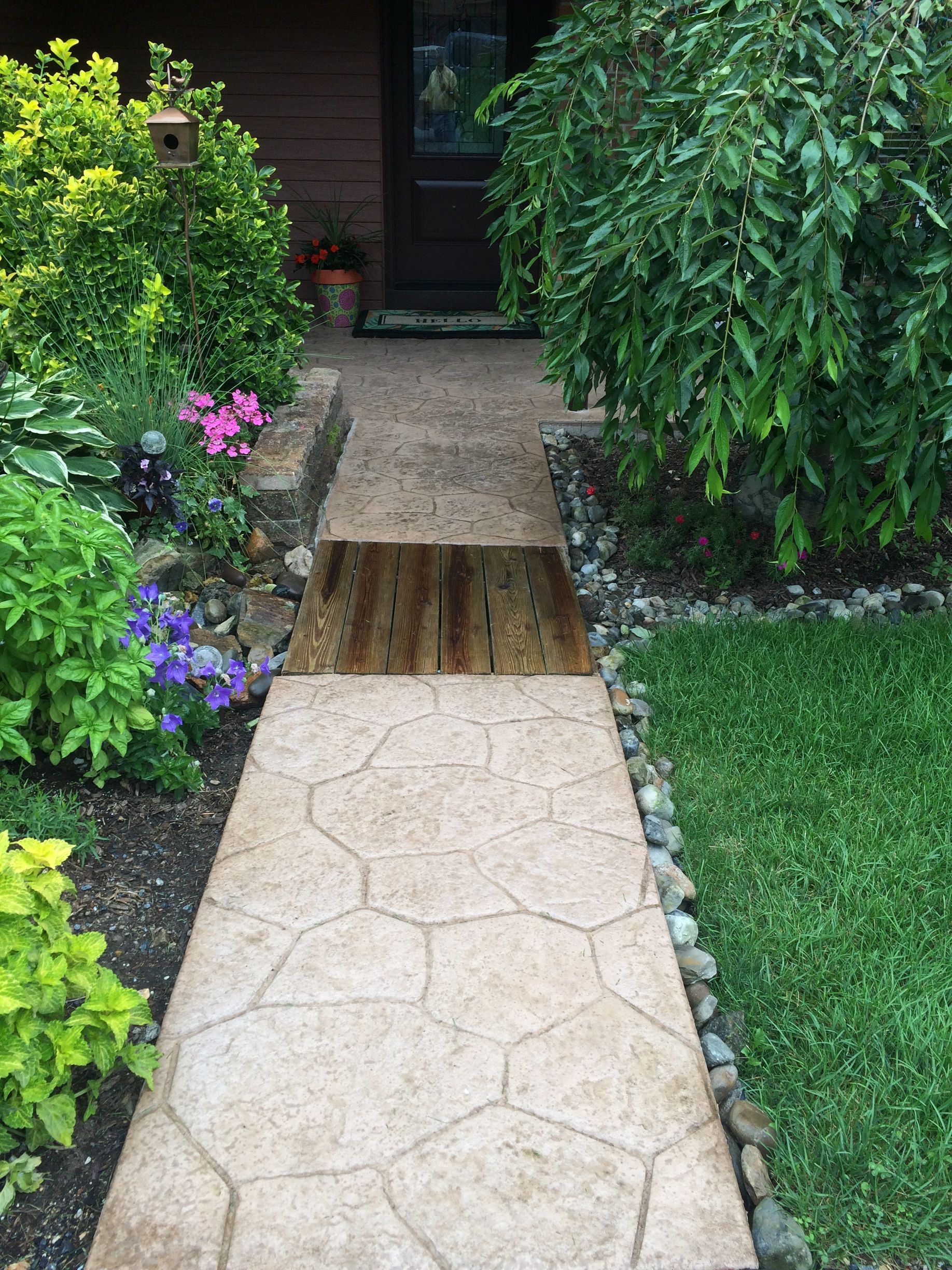 a stamped concrete walkway leading to the front door of a house surrounded by trees and flowers .