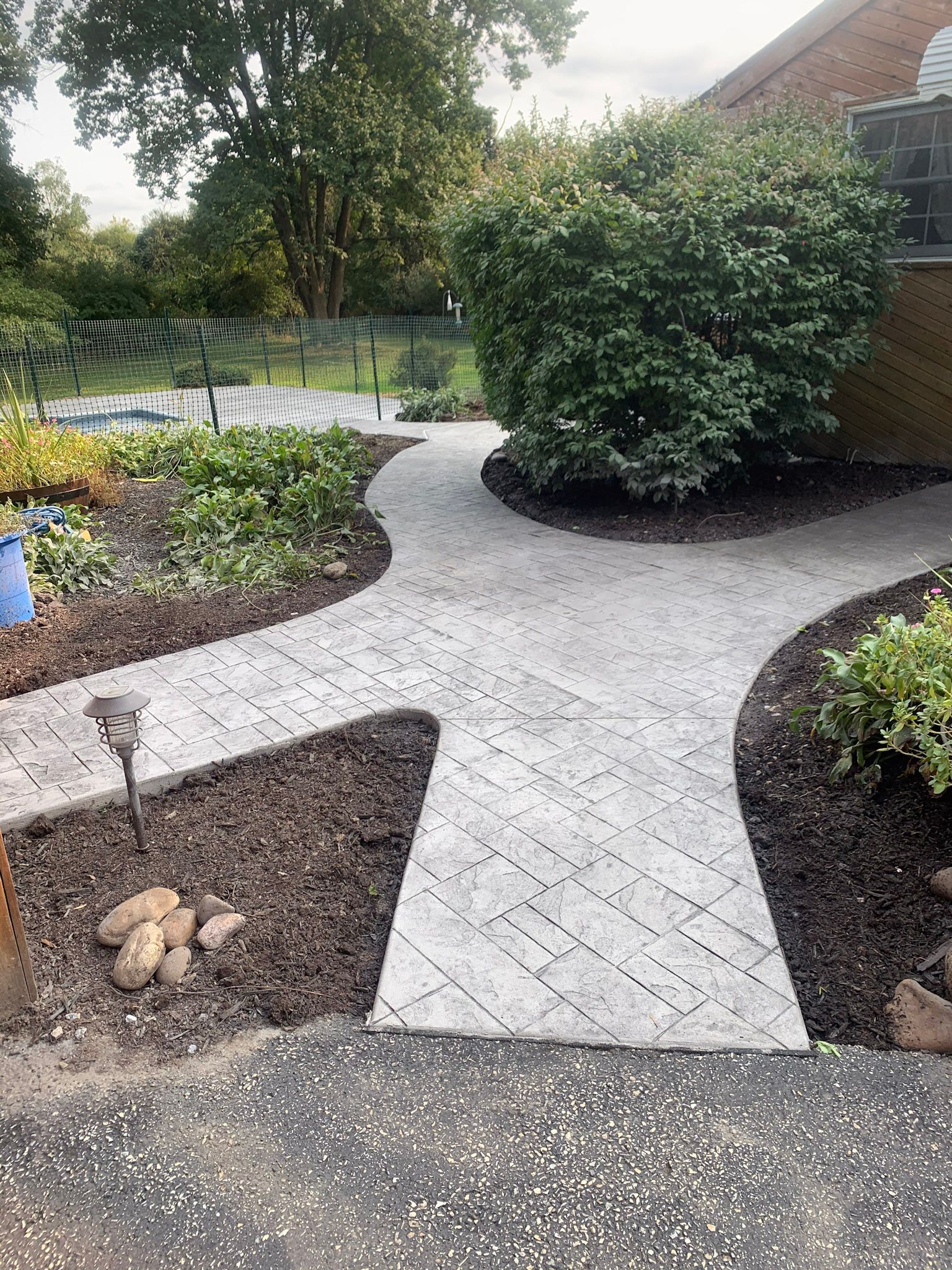 a  stamped concrete brick-like walkway leading to a house surrounded by trees and bushes .