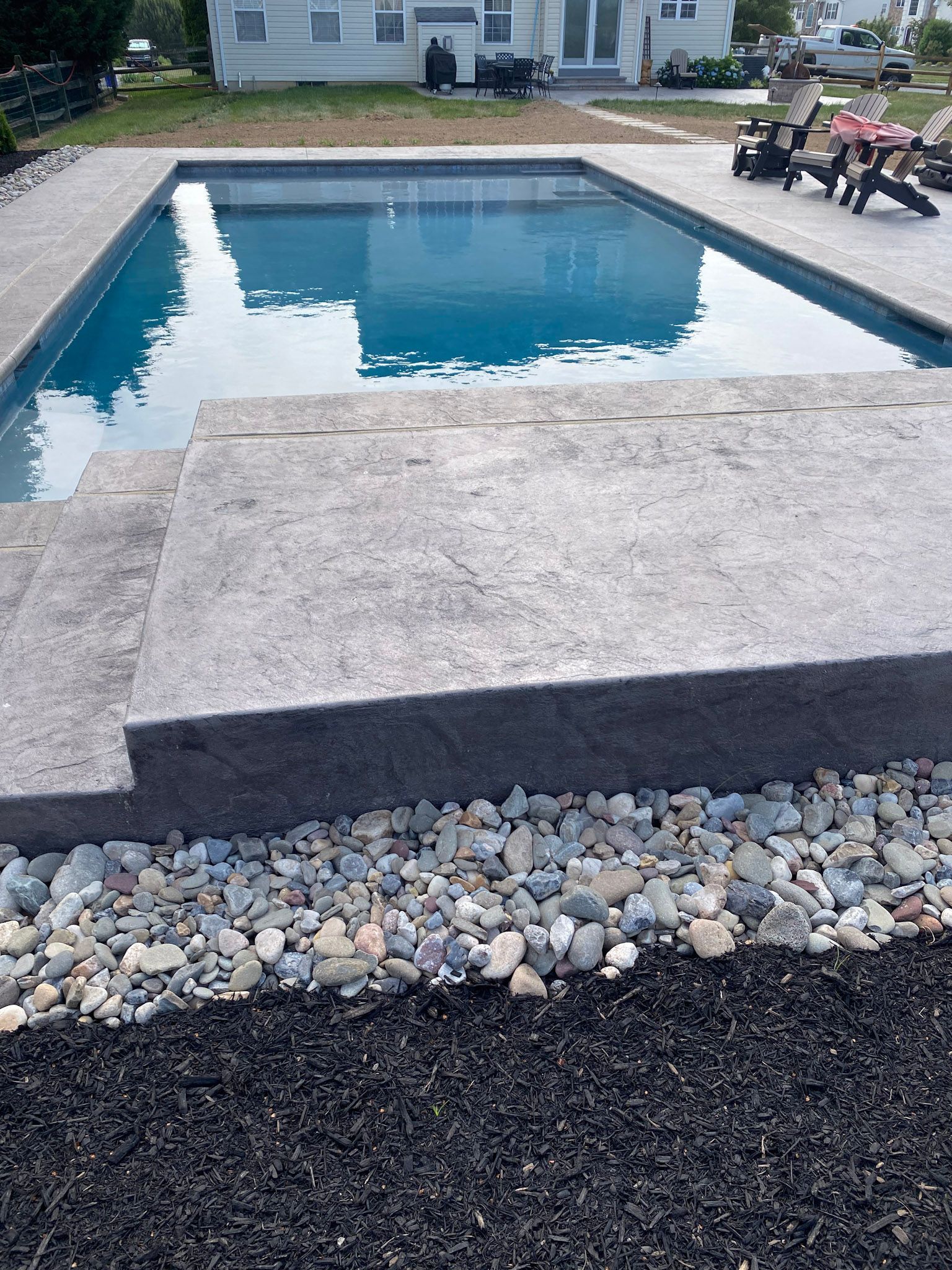 a large  stamped concrete  swimming pool deck is surrounded by rocks and a house in the background
