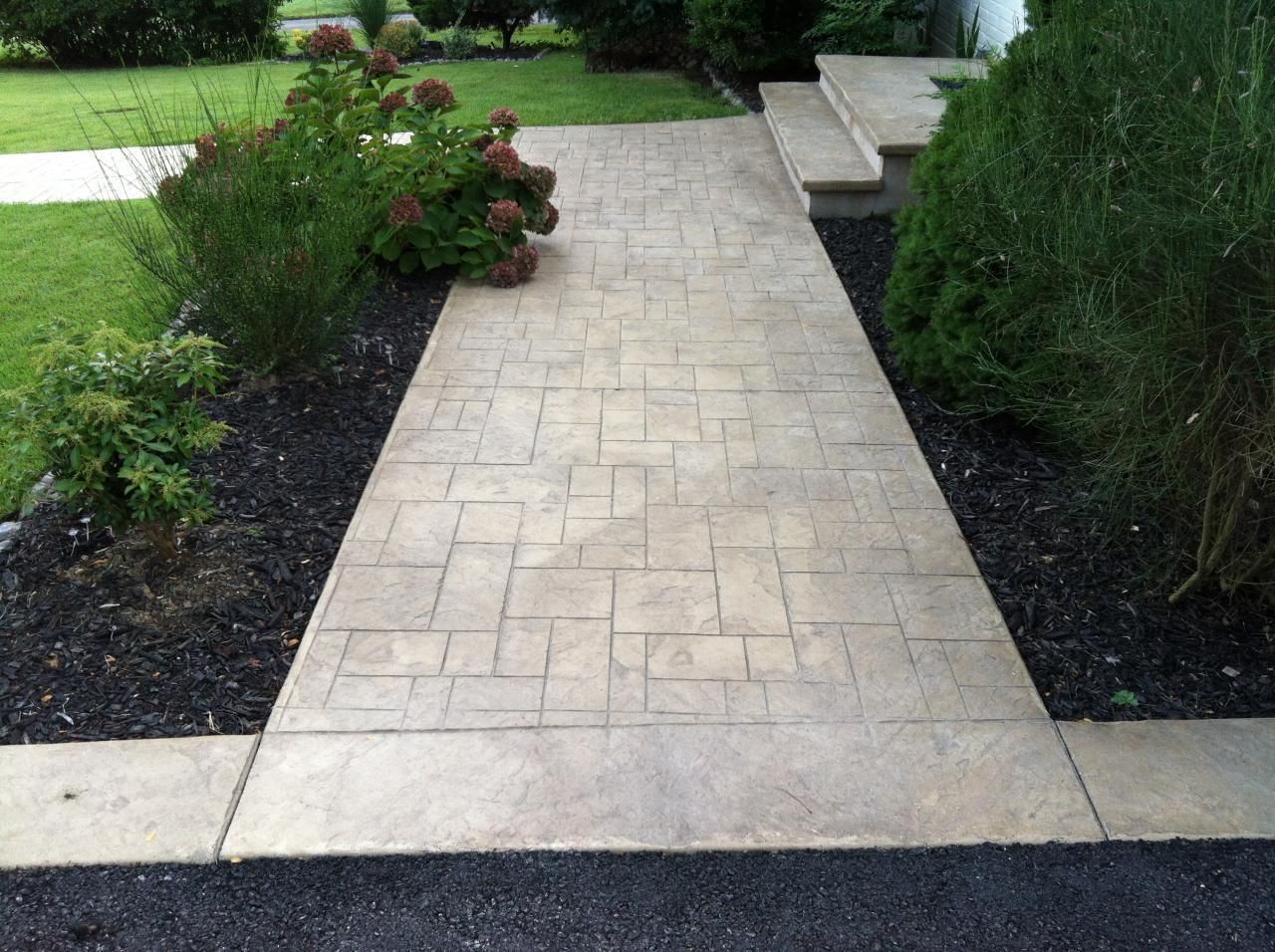 a stamped concrete walkway leading to a house surrounded by trees and bushes