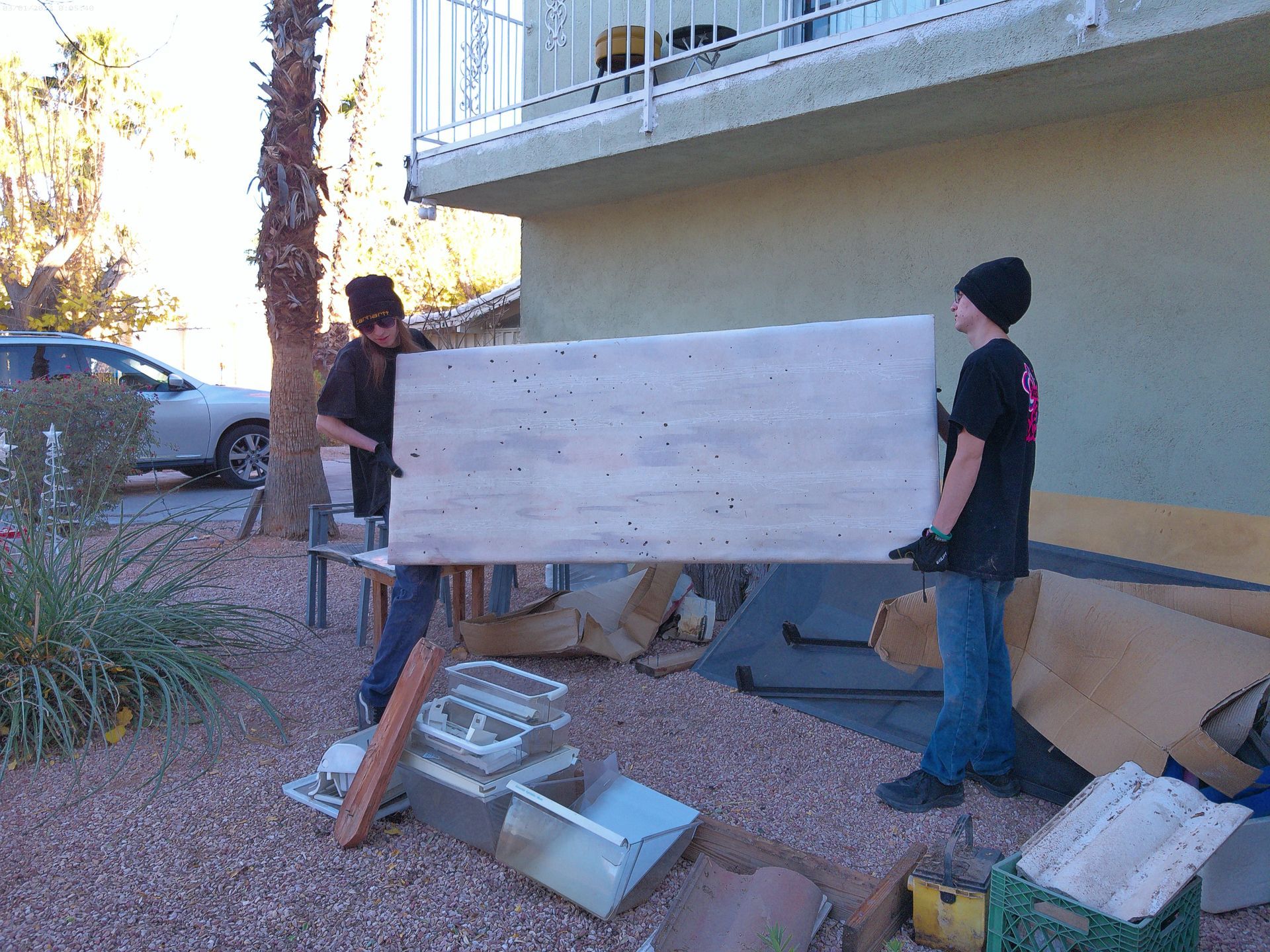Two men are carrying a large piece of wood in front of a building. Removing junk from a yard.