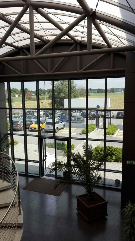 Atrio interno con ampia parete vetrata affacciata su un parcheggio e sull'acqua, con travi in ​​acciaio a vista nella parte superiore.