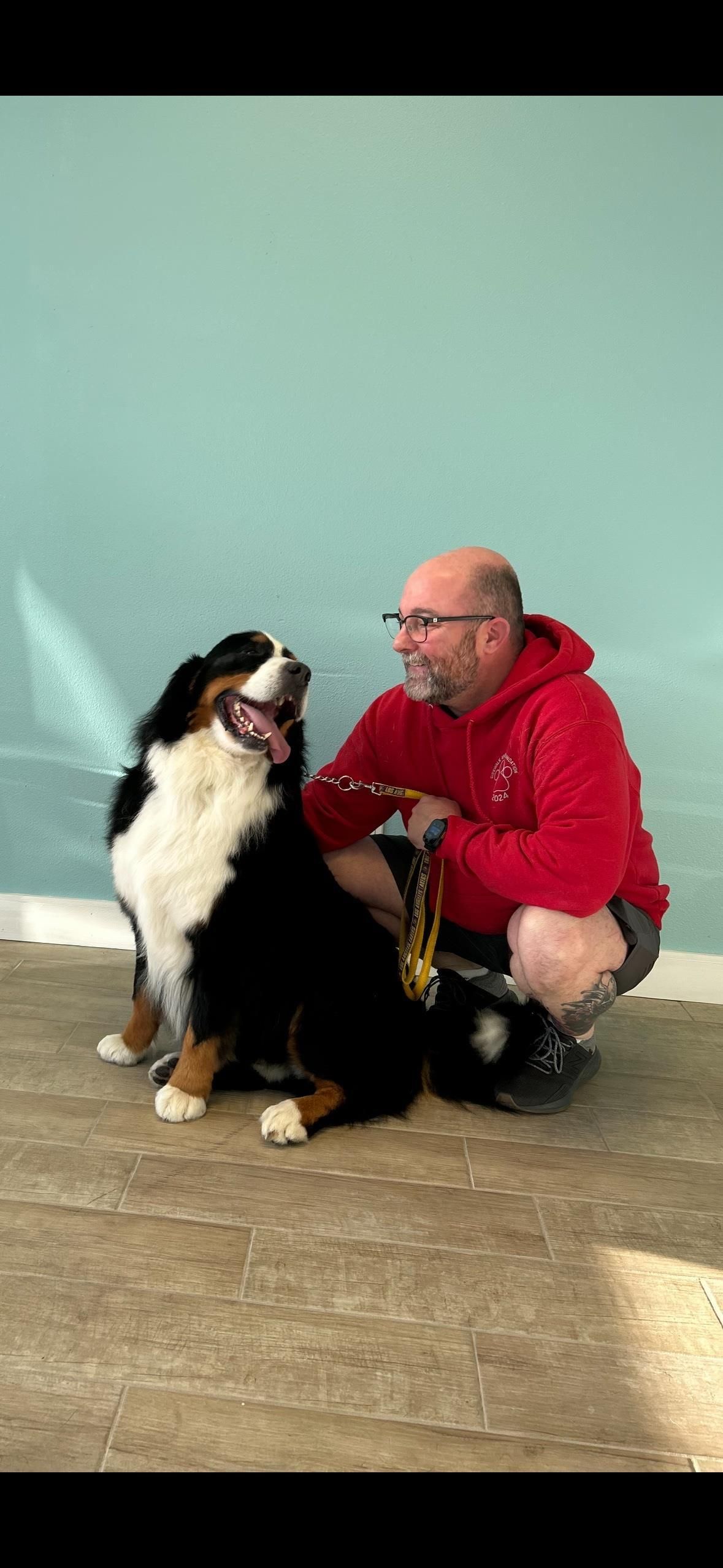 A man in red crouches next to a Bernese mountain dog, both looking at each other. Light blue wall background.