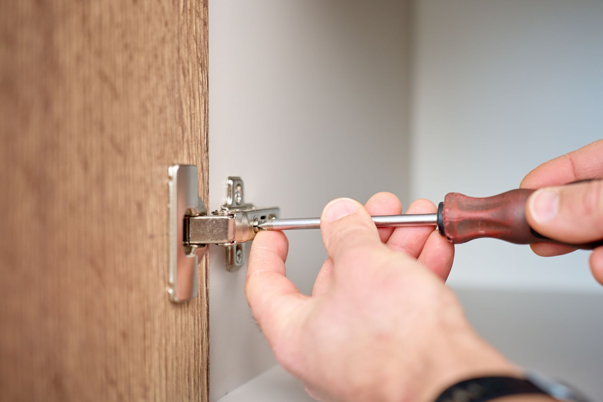 Hands adjusting a metal cabinet hinge with a screwdriver on a wooden door panel