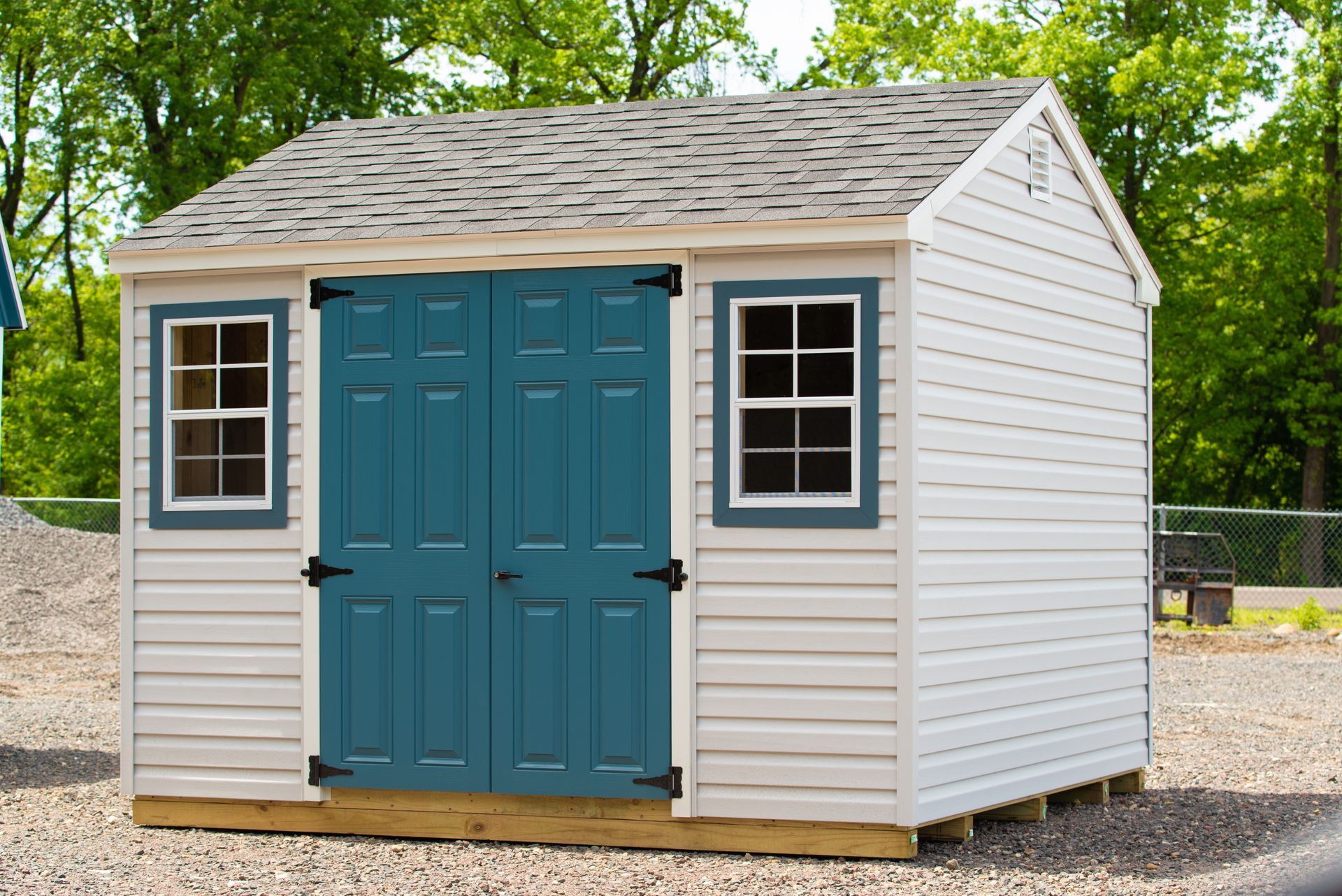 Small white shed with teal double doors and black-trimmed windows on a gravel lot, surrounded by trees
