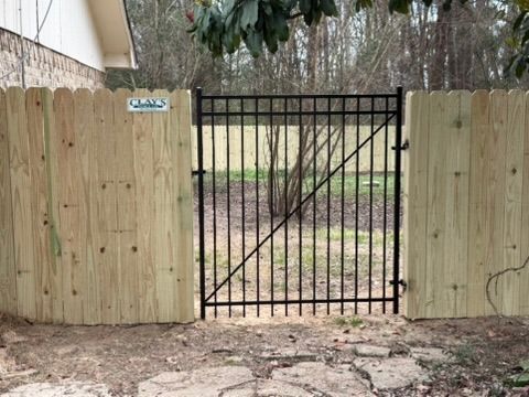 A black metal custom gate set between two wooden picket fences on a gravel path in Tyler, Texas.
