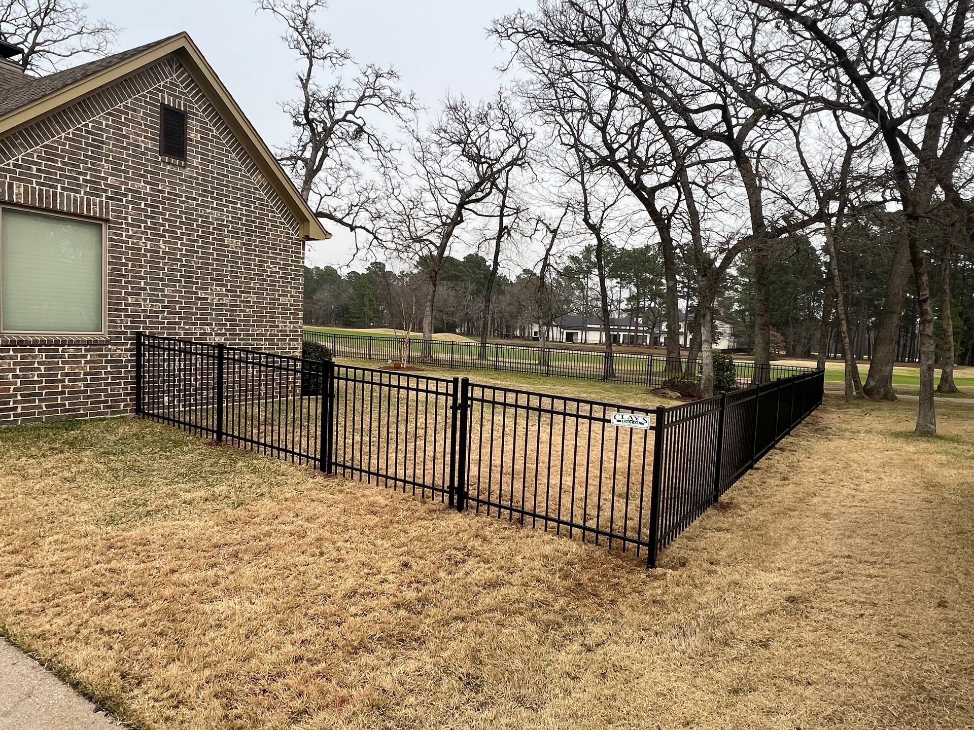 Black metal fence surrounds a yard with brown grass next to a brick house and bare trees.