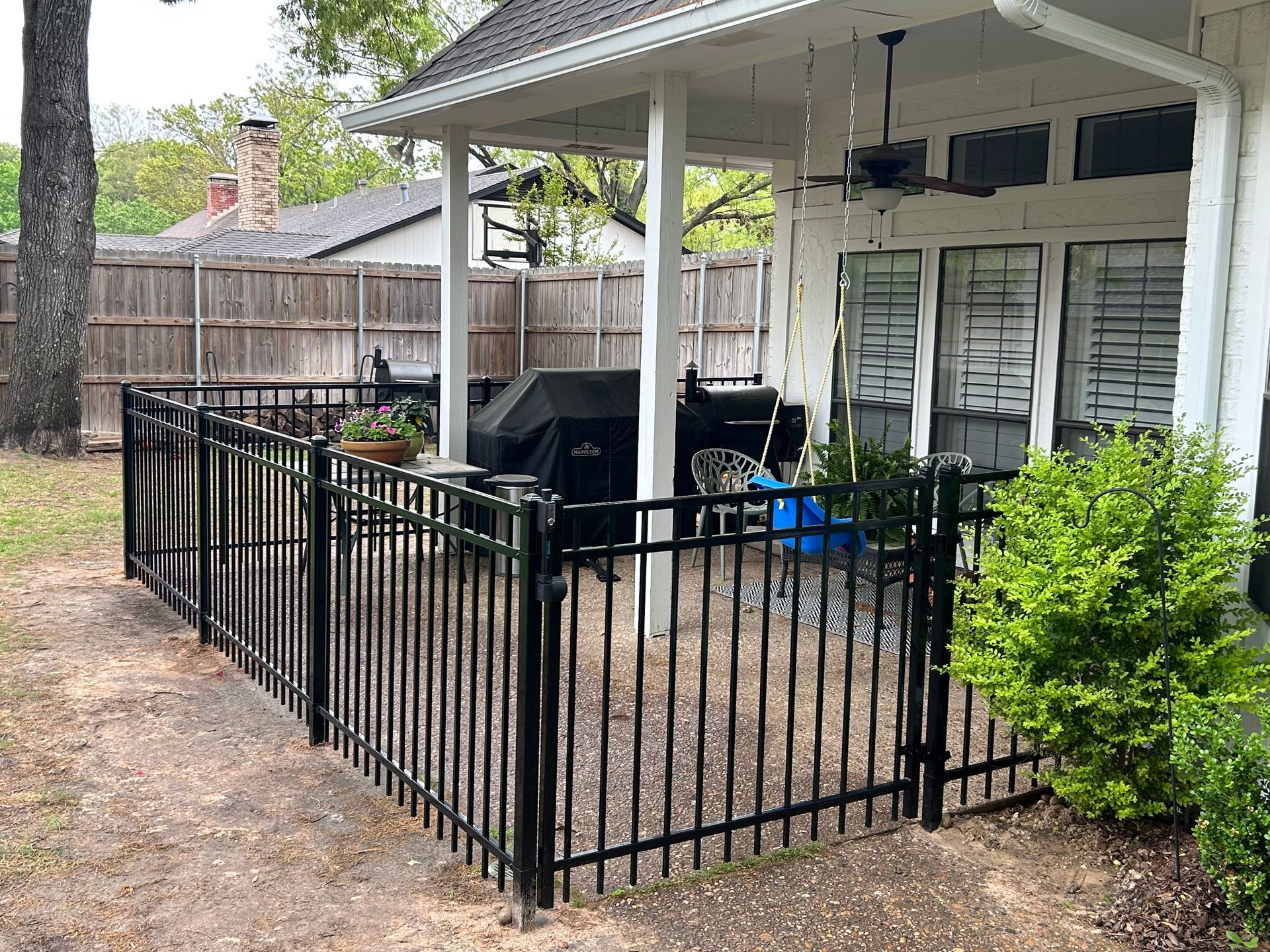 Black wrought-iron fence surrounds a patio with a grill and seating; white house in the background.