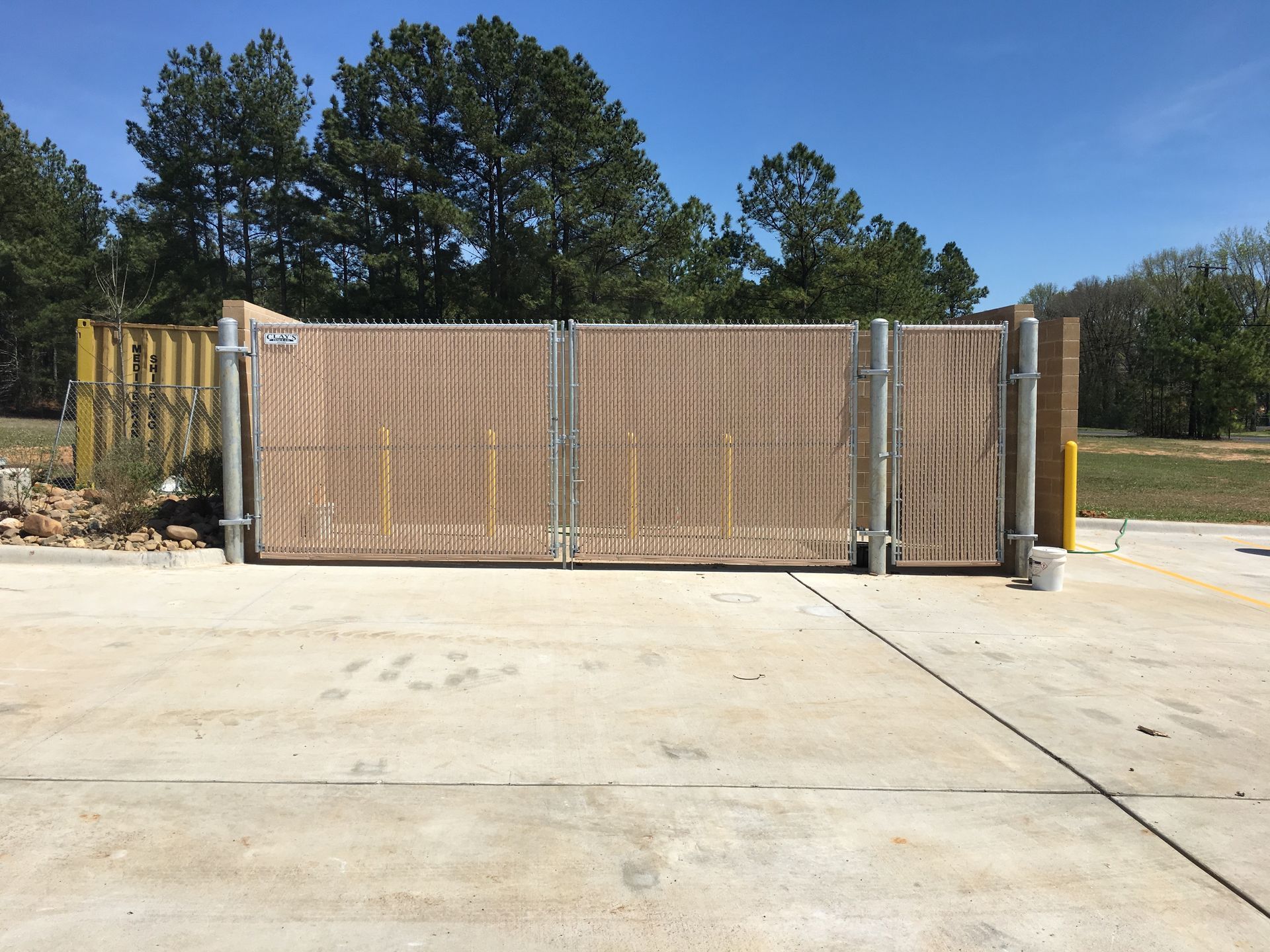 Chain-link fence with tan slats encloses a trash area on a concrete surface. Trees and blue sky in background.