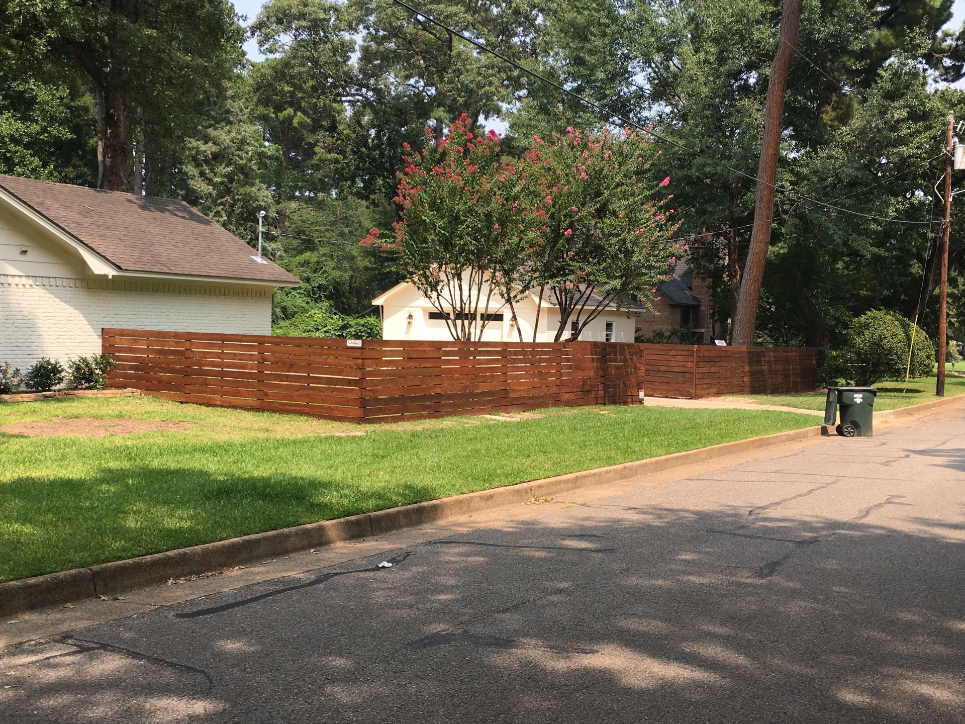 A brown wooden fence surrounds a house, with green grass and a street in front.
