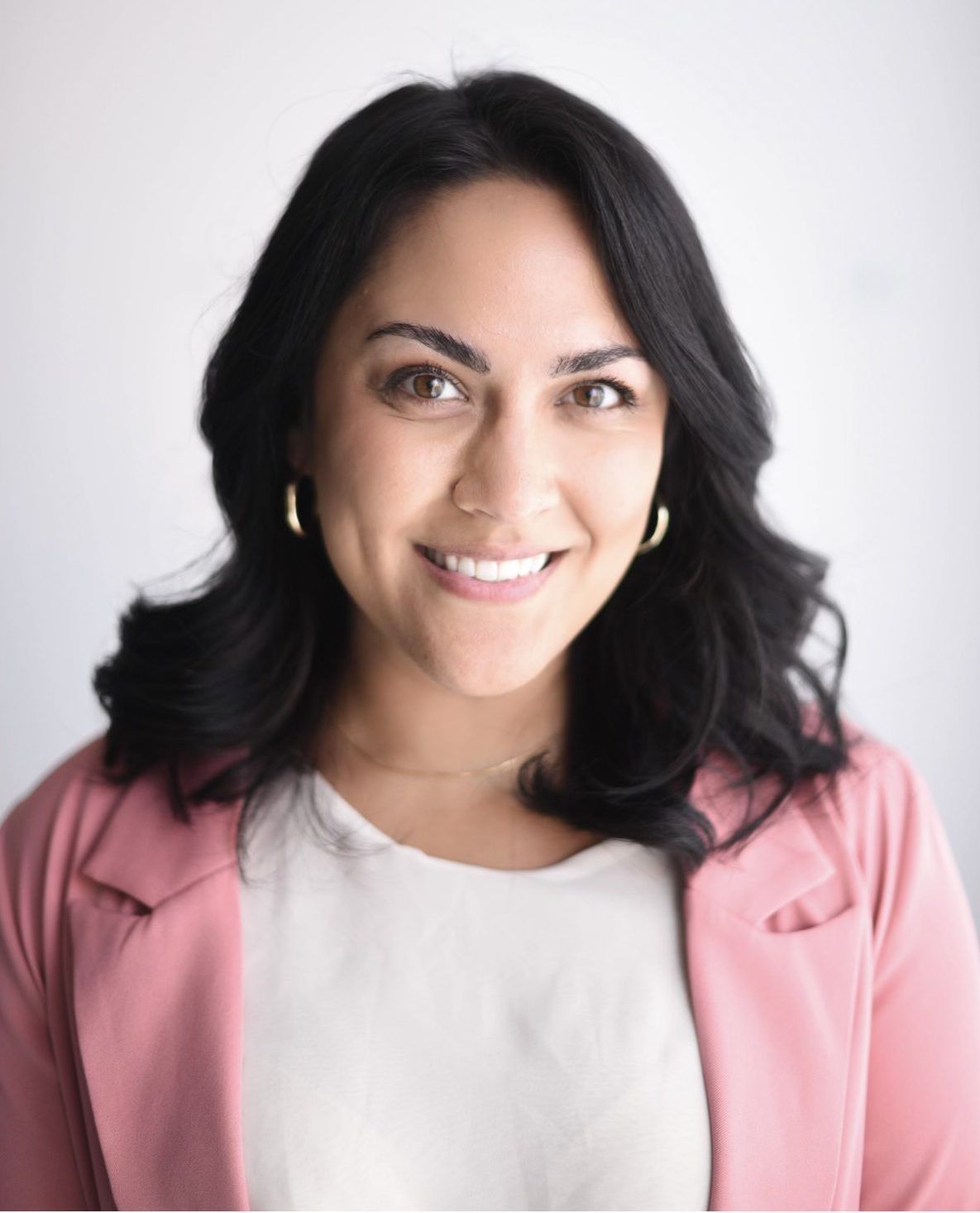 A woman wearing a pink jacket and earrings is smiling for the camera.