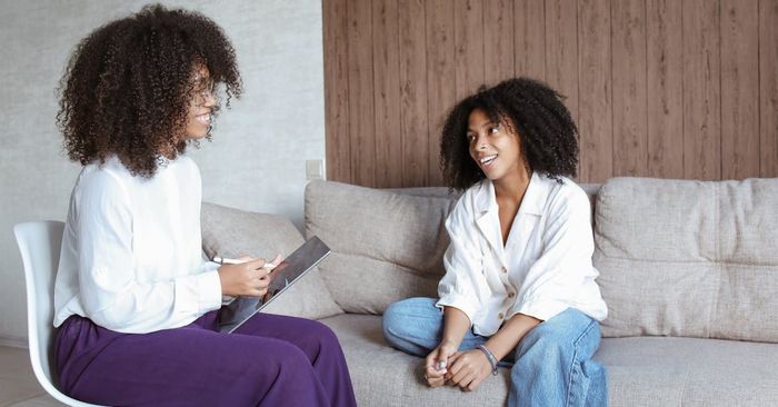 Two women are sitting on a couch talking to each other.