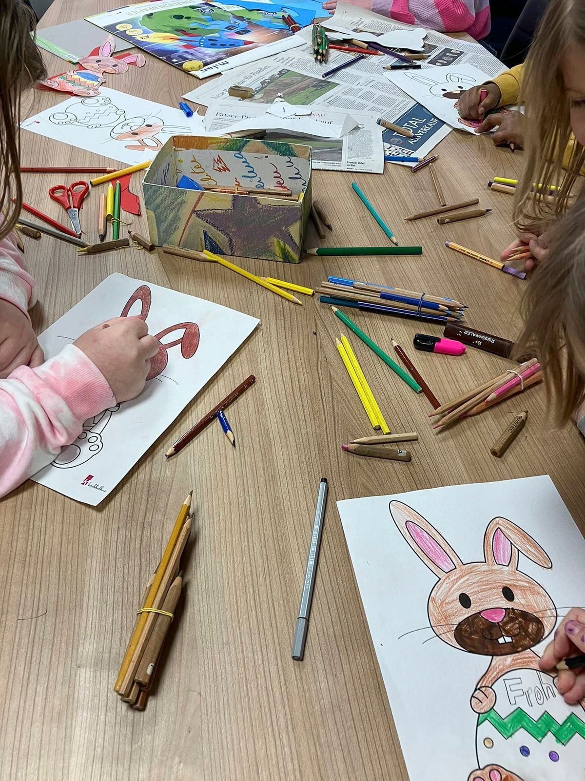 Ein Tisch mit vielen Malstiften, zu erkennen sind mehrere Kinder, die Osterhasen malen