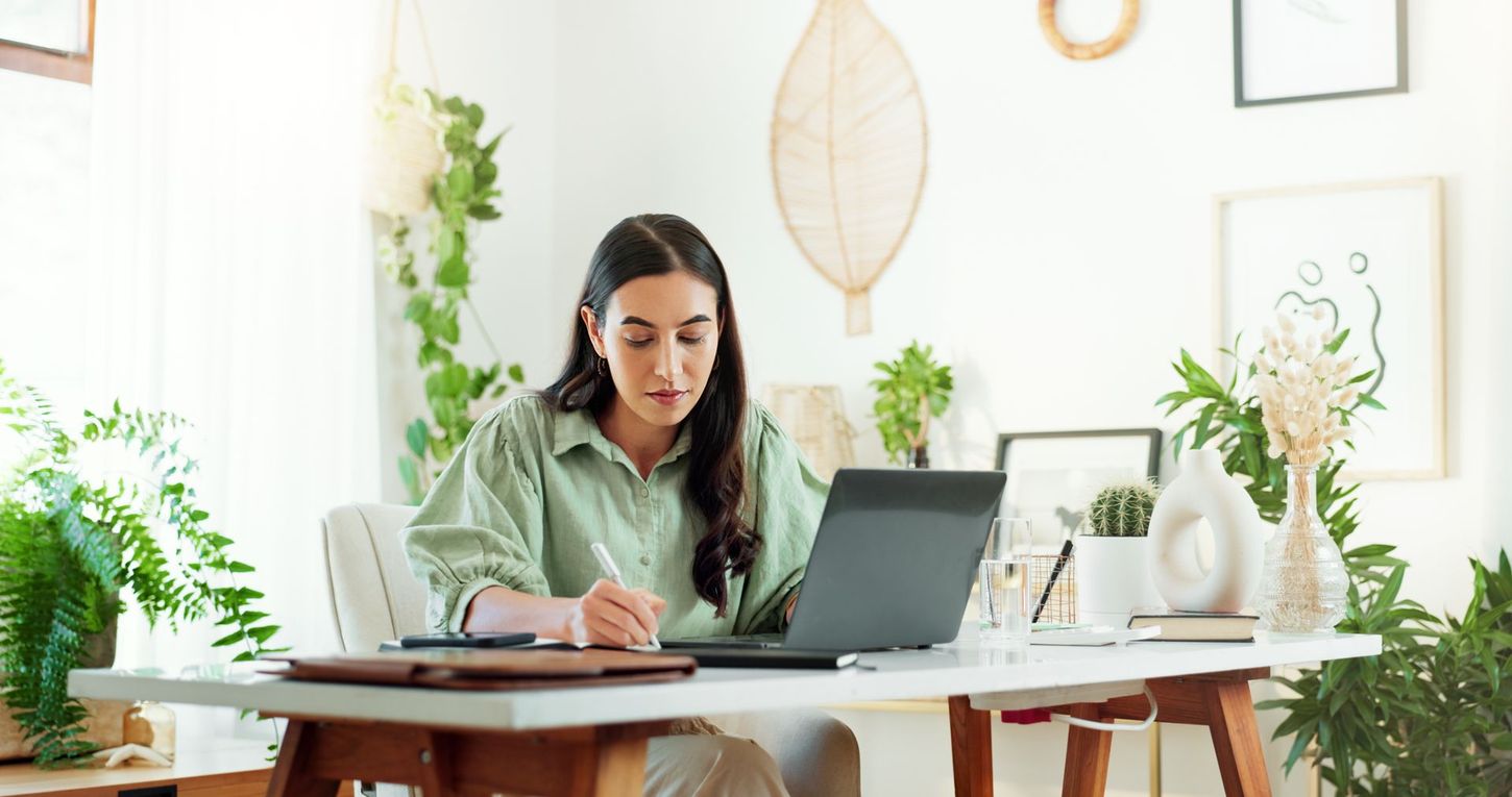 Woman working on laptop at a white desk surrounded by plants in a bright room.