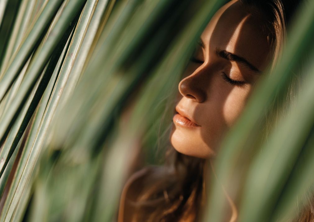 Volto di donna esposto alla luce del sole, occhi chiusi, incorniciato da foglie verdi.