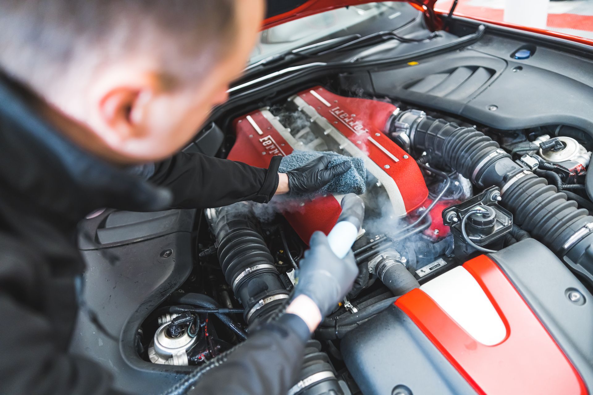 A person wearing gloves steam cleans the red engine bay of a car.