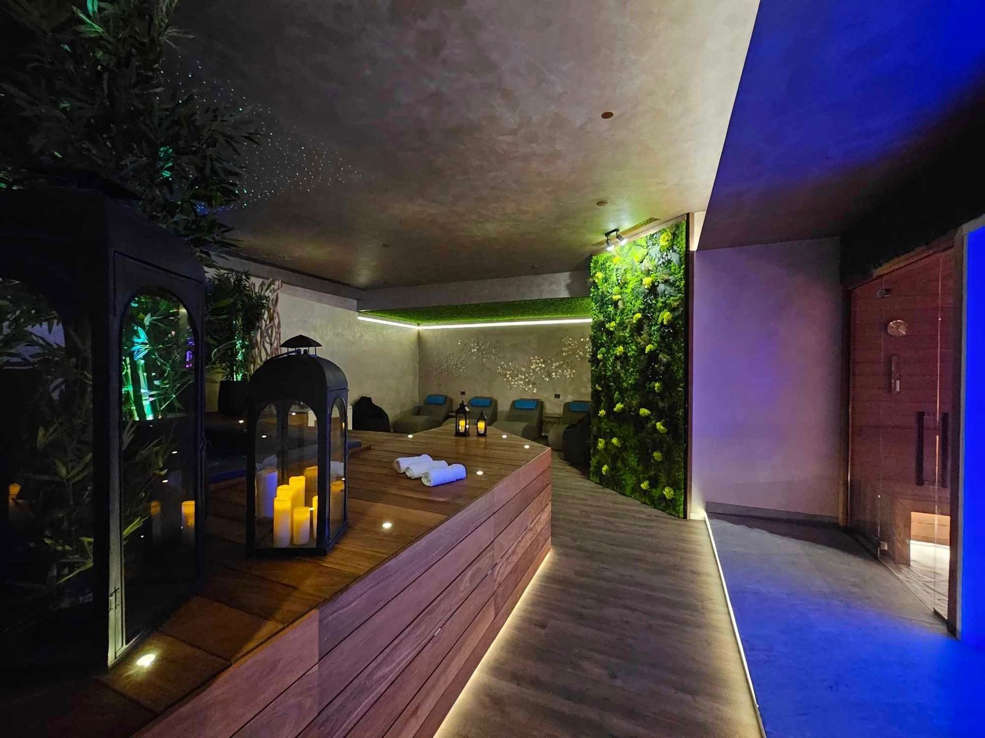 Interno della spa con bancone in legno e candele. Lungo corridoio con sedute e una parete di piante.
