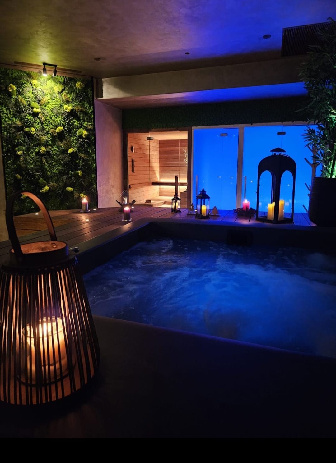 Accogliente sala spa con vasca idromassaggio, illuminata da lanterne e candele, dotata di sauna e parete verde.