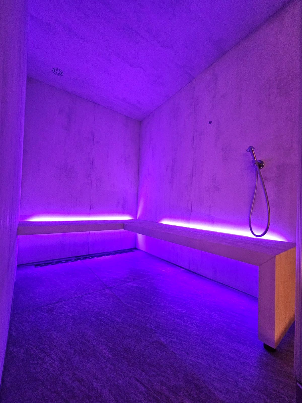 Doccia termale illuminata di viola. Pareti in cemento, panca con luce di fondo, soffione e pavimento scuro.